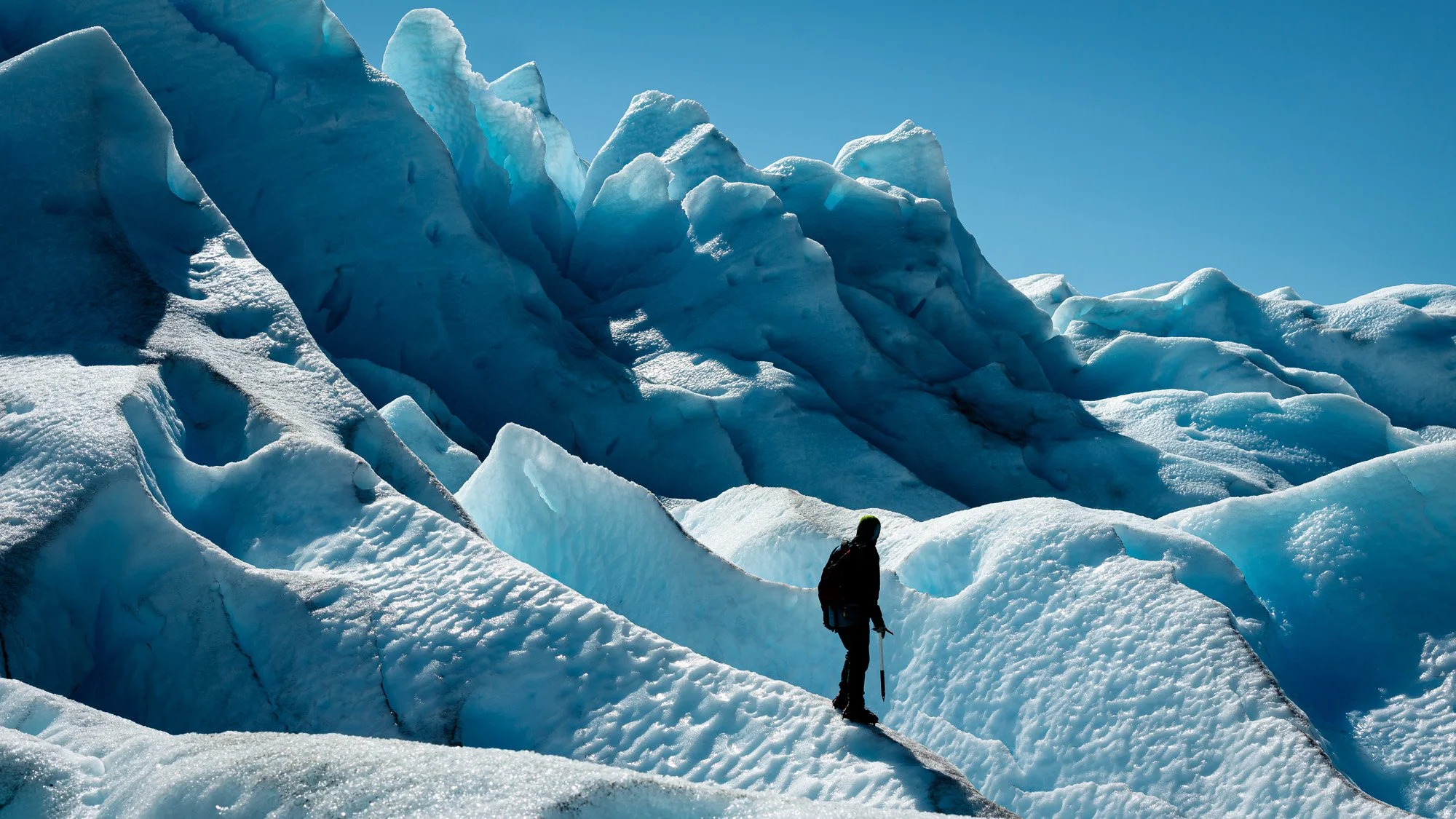 A lone adventurer navigates the deep blue crevasses of the Perito Moreno Glacier in Patagonia, Argentina. The high figure-to-ground contrast of the human silhouette against the ice establishes immediate narrative context and scale.