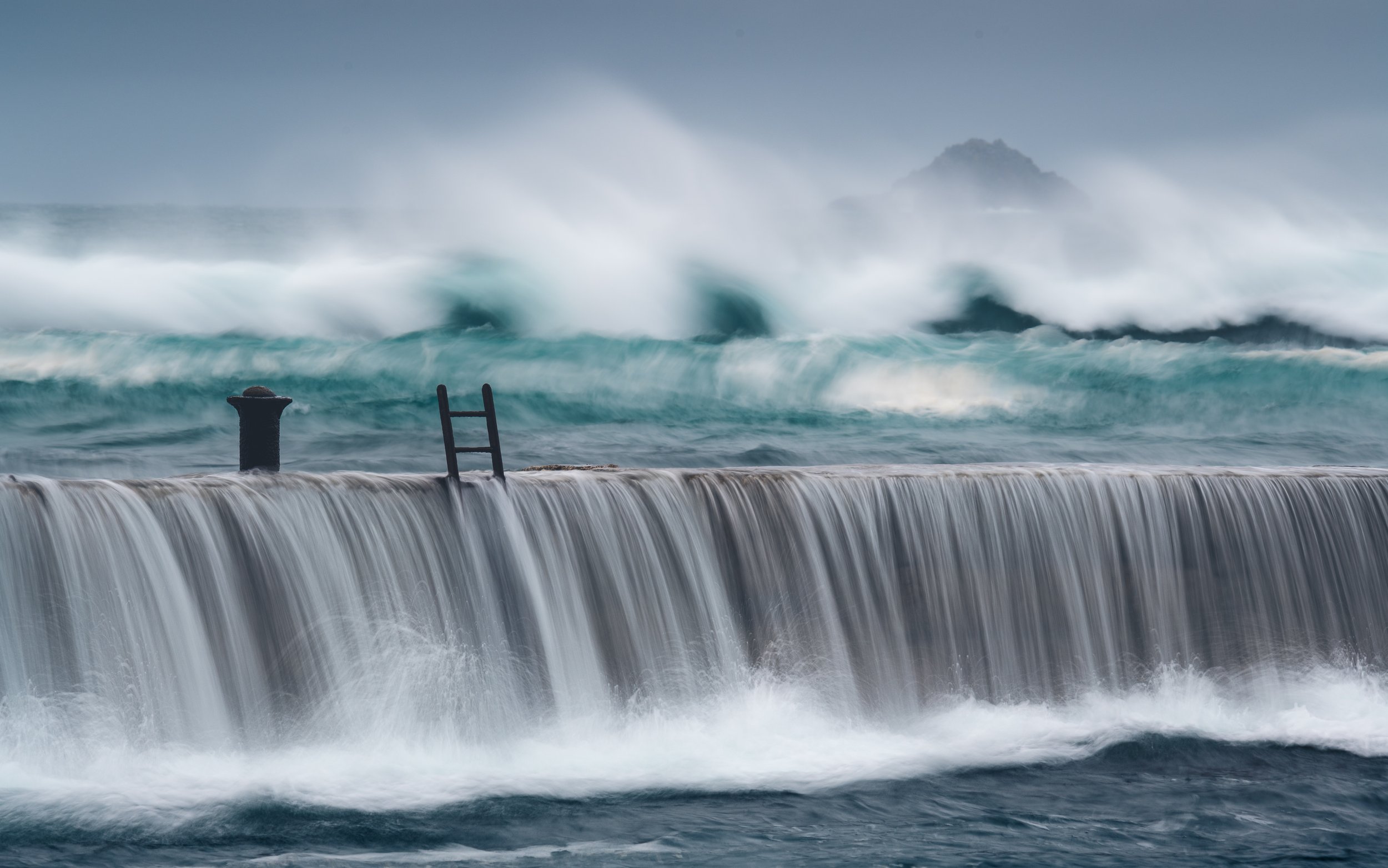 Rough ocean waves crashing over a concrete barrier with a ladder and a post, engulfed in spray and mist, with a distant island in the background