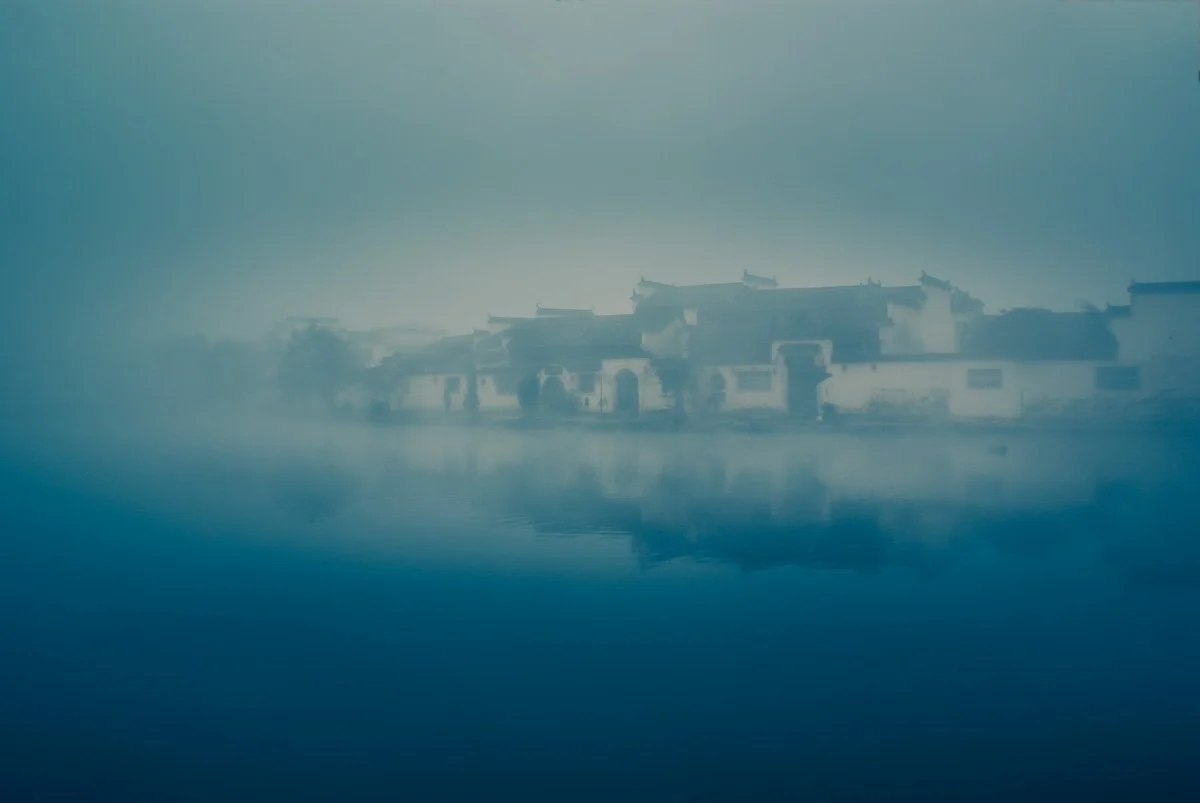 The historic whitewashed buildings of an ancient Chinese village in Anhui submerged in deep blue morning fog and reflected in still water.
