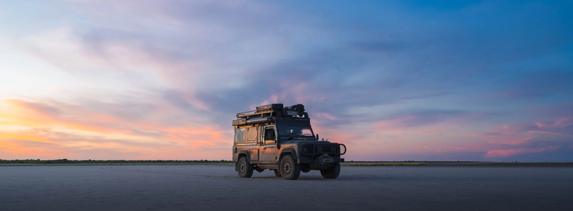 A Land Rover Defender parked on the flat expanse of the Makgadikgadi Pans in Botswana under a twilight sky.
