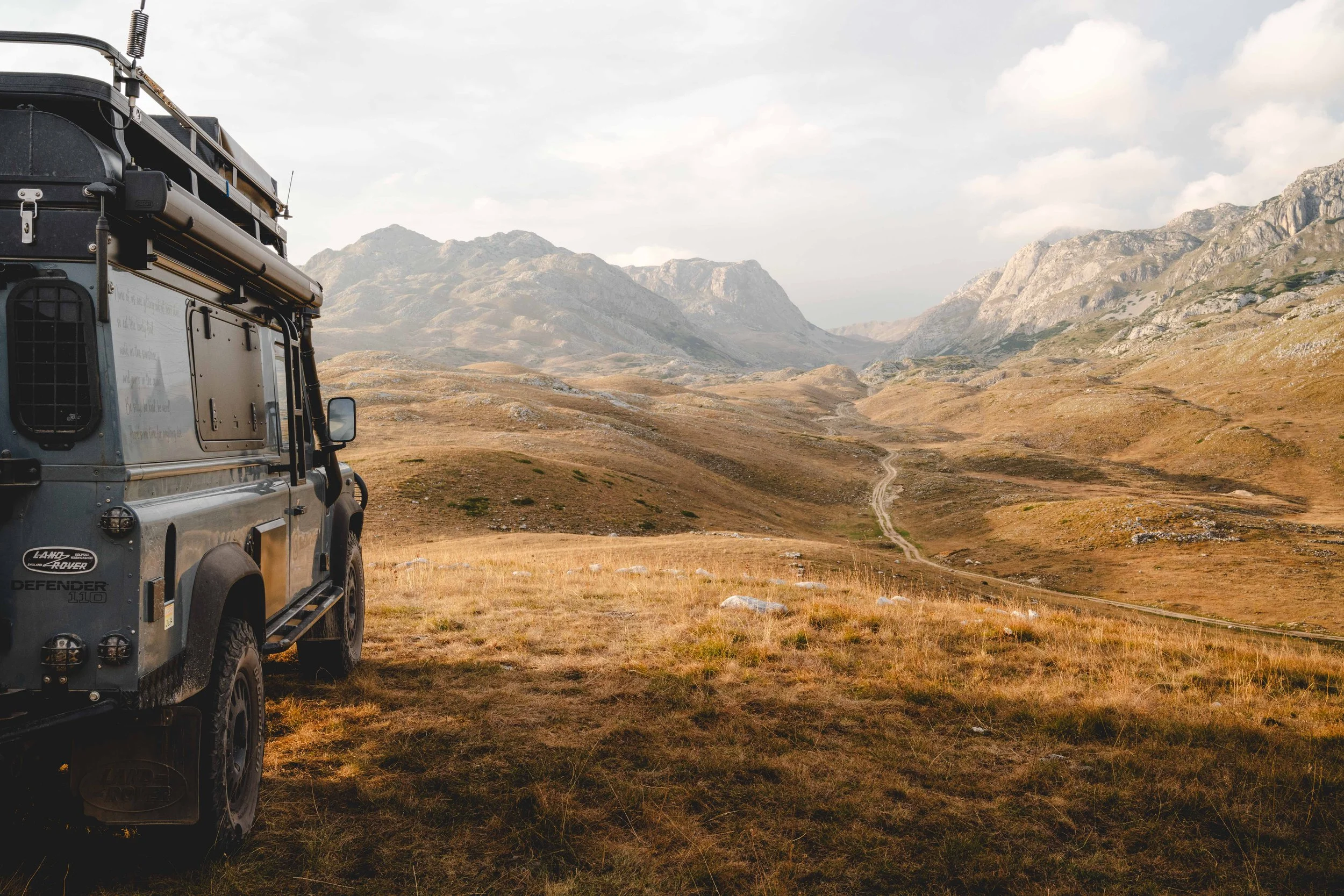 A rugged Land Rover Defender parked on a grassy hillside overlooking a valley with winding dirt roads and mountains in the background.