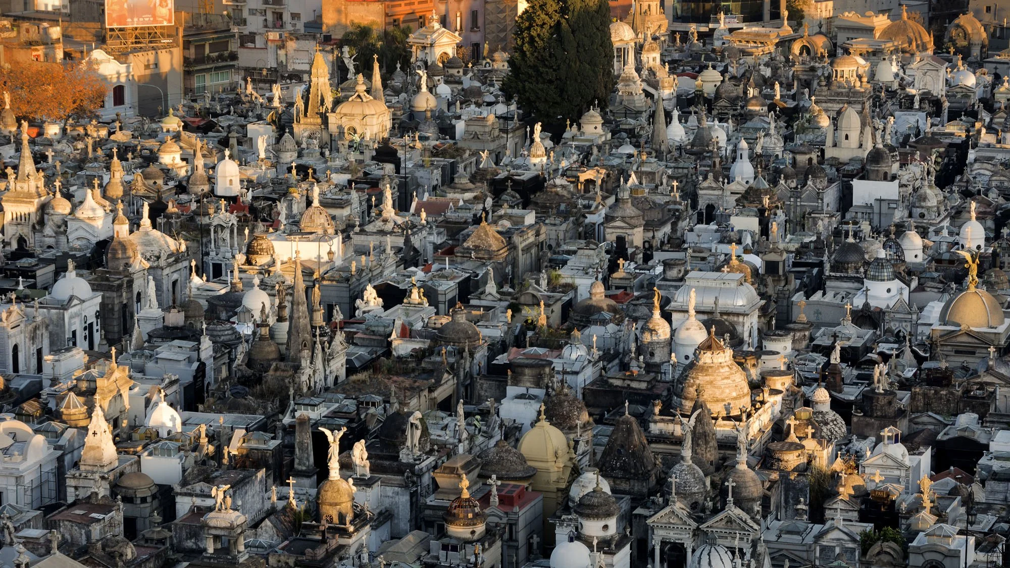 A high-angle view overlooking the dense stone mausoleums, domes, and statues of Recoleta Cemetery in Buenos Aires, bathed in warm morning golden-hour light