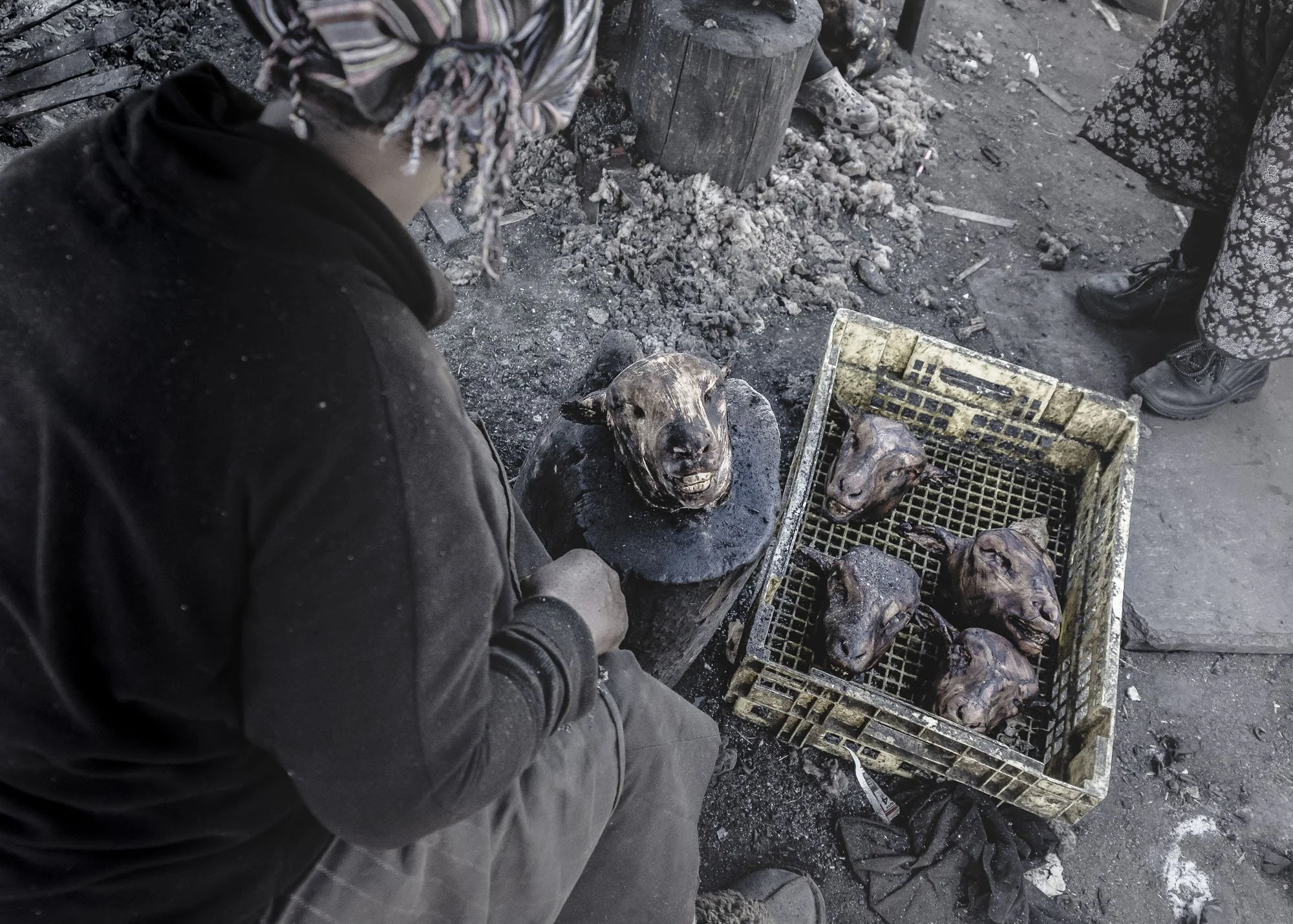 An overhead documentary view of a woman preparing singed sheep heads over charcoal embers in a gritty market in Langa Township, South Africa
