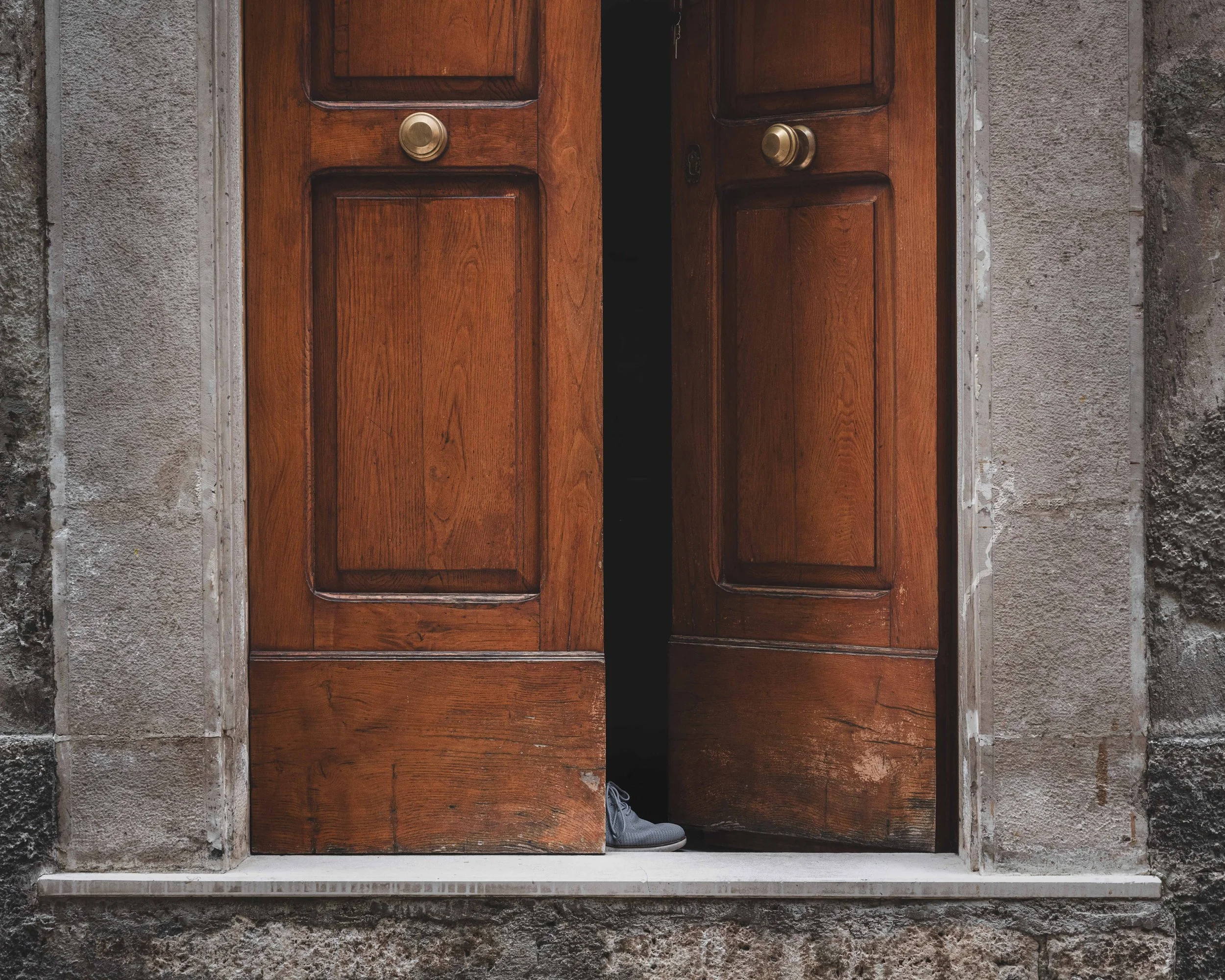 Wooden double doors with brass doorknobs, slightly open, revealing a sneaker inside the dark doorway, surrounded by stone walls.
