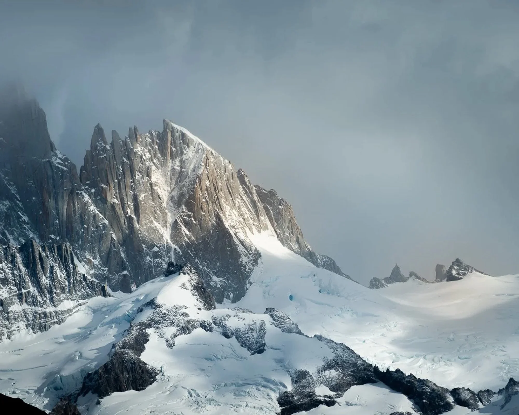 The jagged granite peaks of the Fitz Roy range in Patagonia, Argentina, partially obscured by heavy clouds. The dramatic side-lighting on the rock face creates a high-contrast attention signal, emphasizing the rugged texture of the mountain.