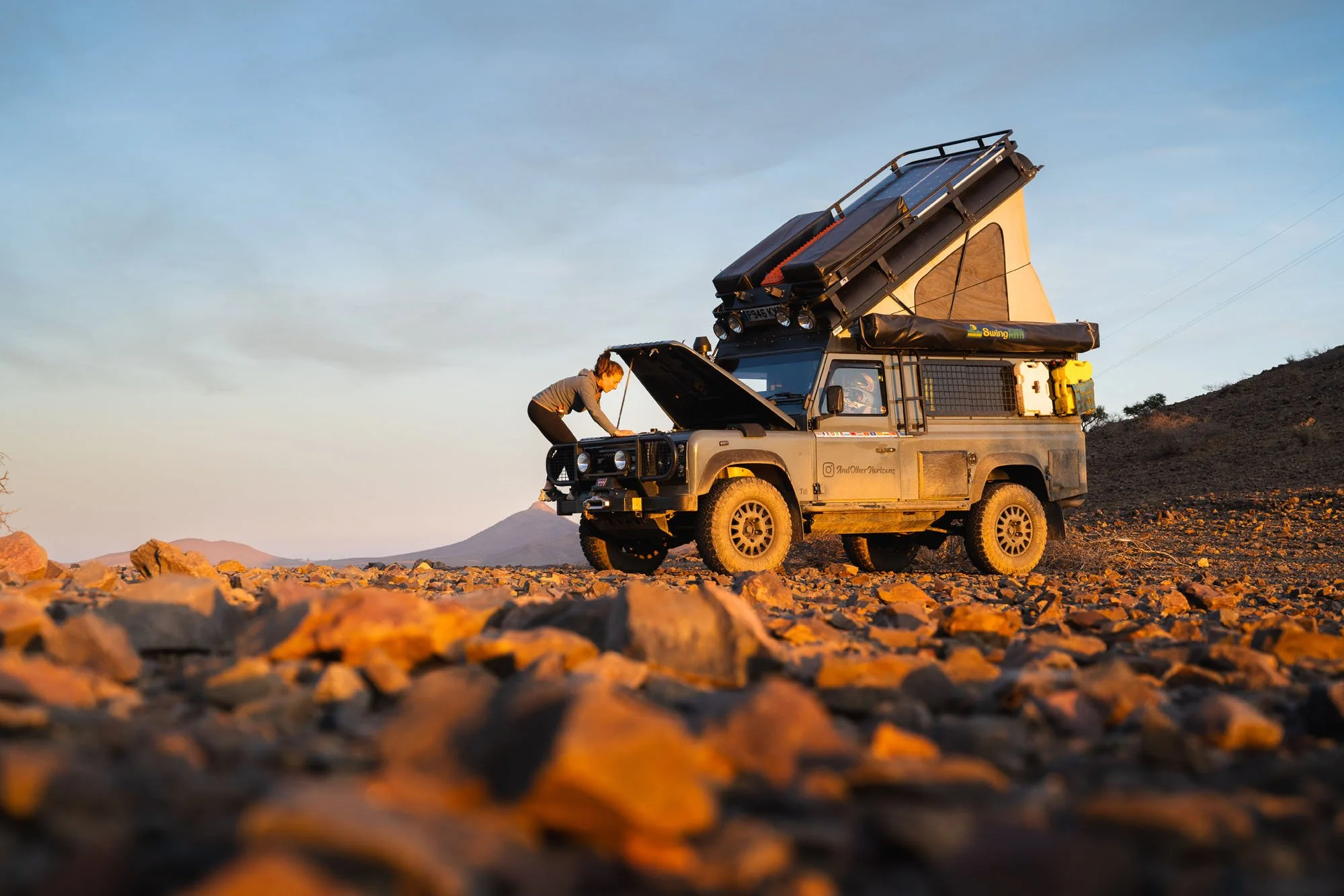 A traveler inspecting the open engine bay of an overland Land Rover Defender with a roof tent on a rocky mountain plateau at sunset in Morocco.