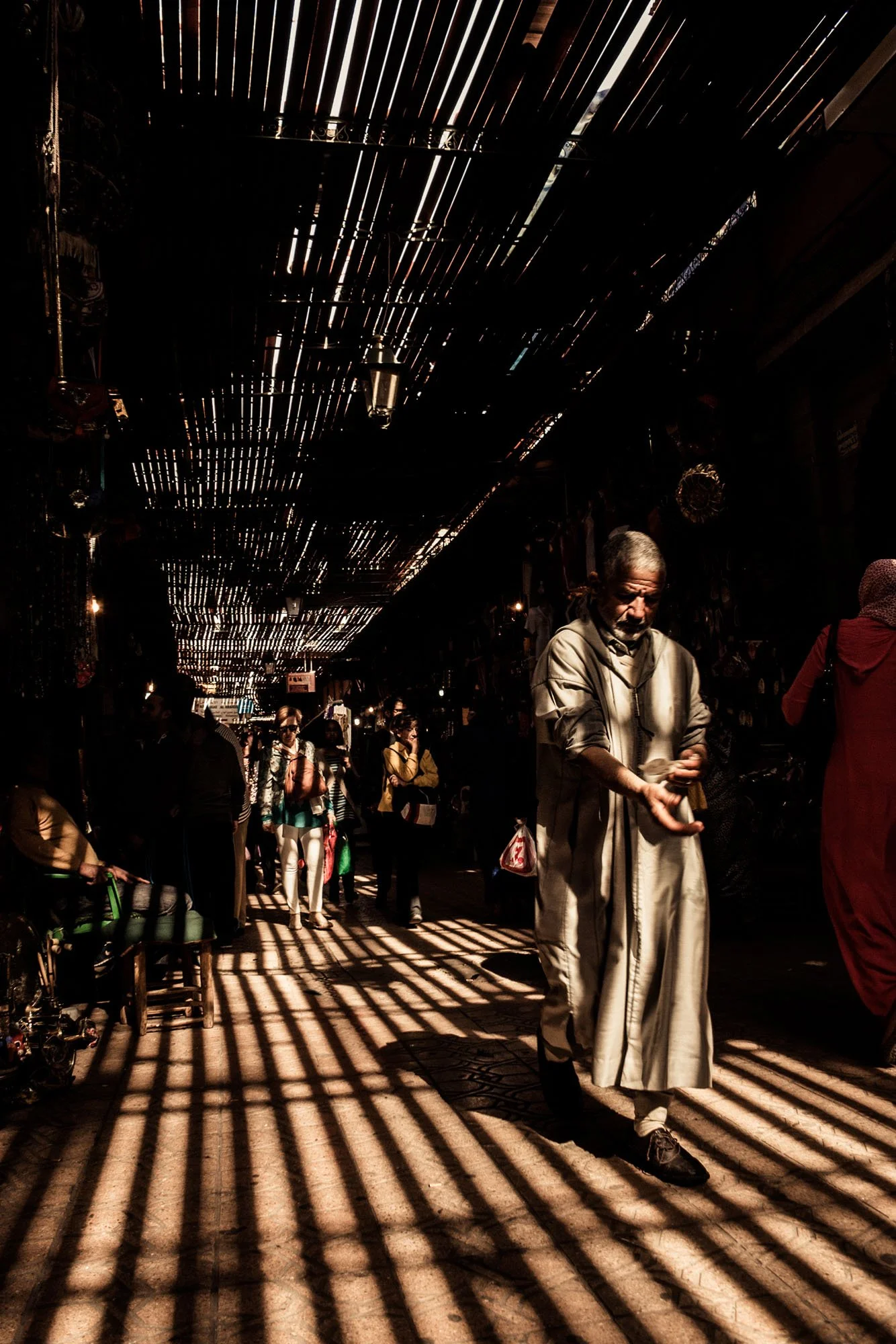 A man in traditional dress walking through a covered alleyway in the Marrakech medina with dramatic striped shadows cast across the floor by a slatted reed ceiling.