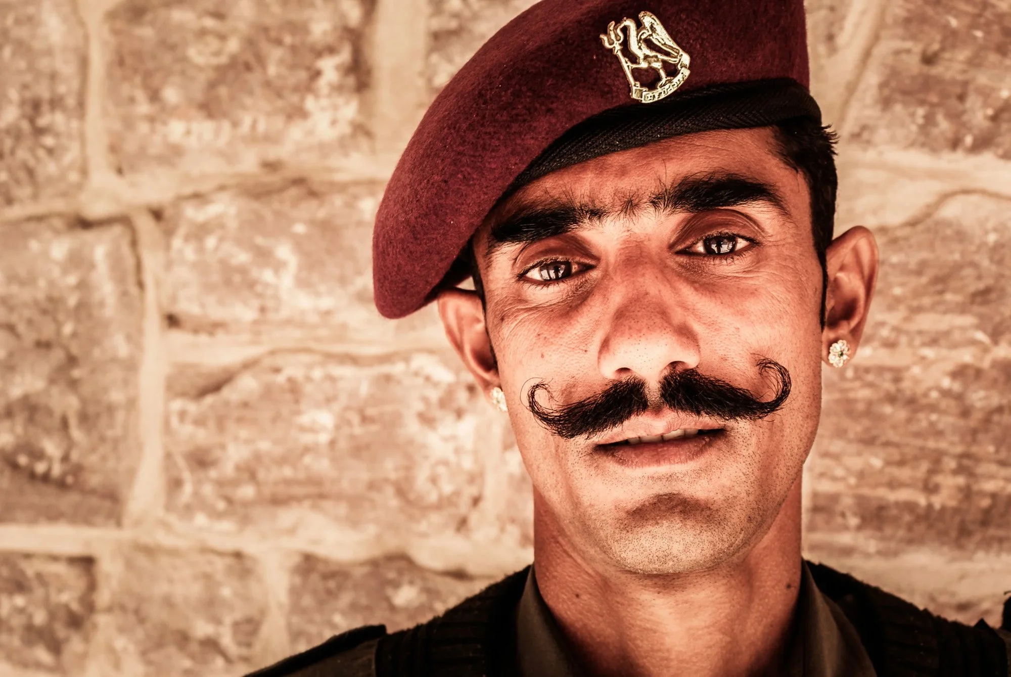 A tight, focused portrait of a guard in Rajasthan, India, wearing a traditional maroon beret and featuring a prominent moustache. His direct, unblinking eye contact acts as a biological tripwire, instantly engaging the viewer’s saliency network.