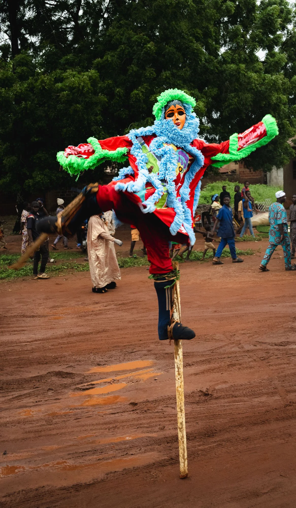 A Moko Jumbie dancer on high wooden stilts leaping in a colorful red and blue fringed costume during a traditional Vodun funeral procession in Benin.