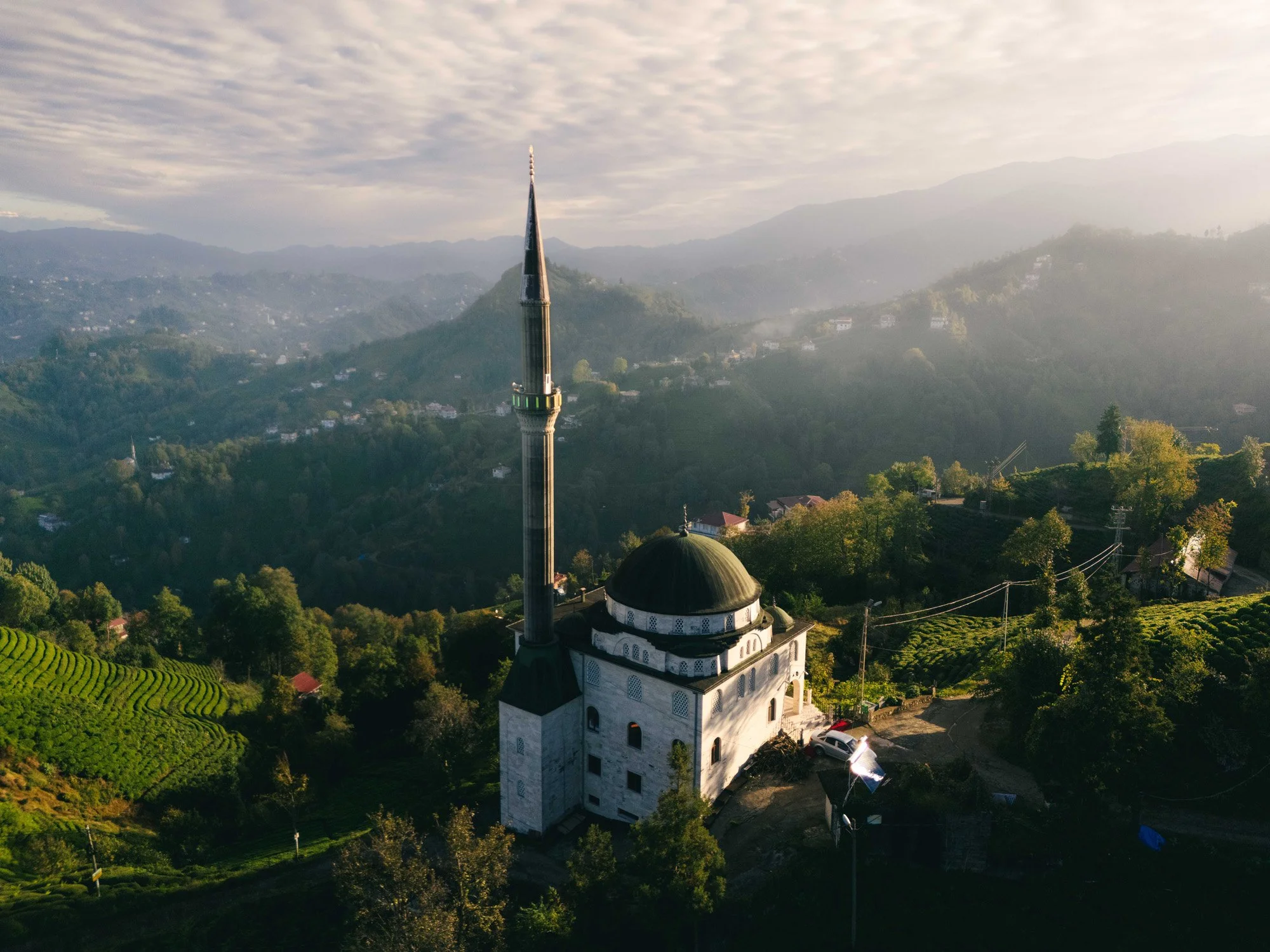 A lonely mosque minaret rises above the mist-kissed tea plantations of Haremtepe, Türkiye. The atmospheric perspective and soft sunrise light create a psychological sense of peace and spiritual energy.