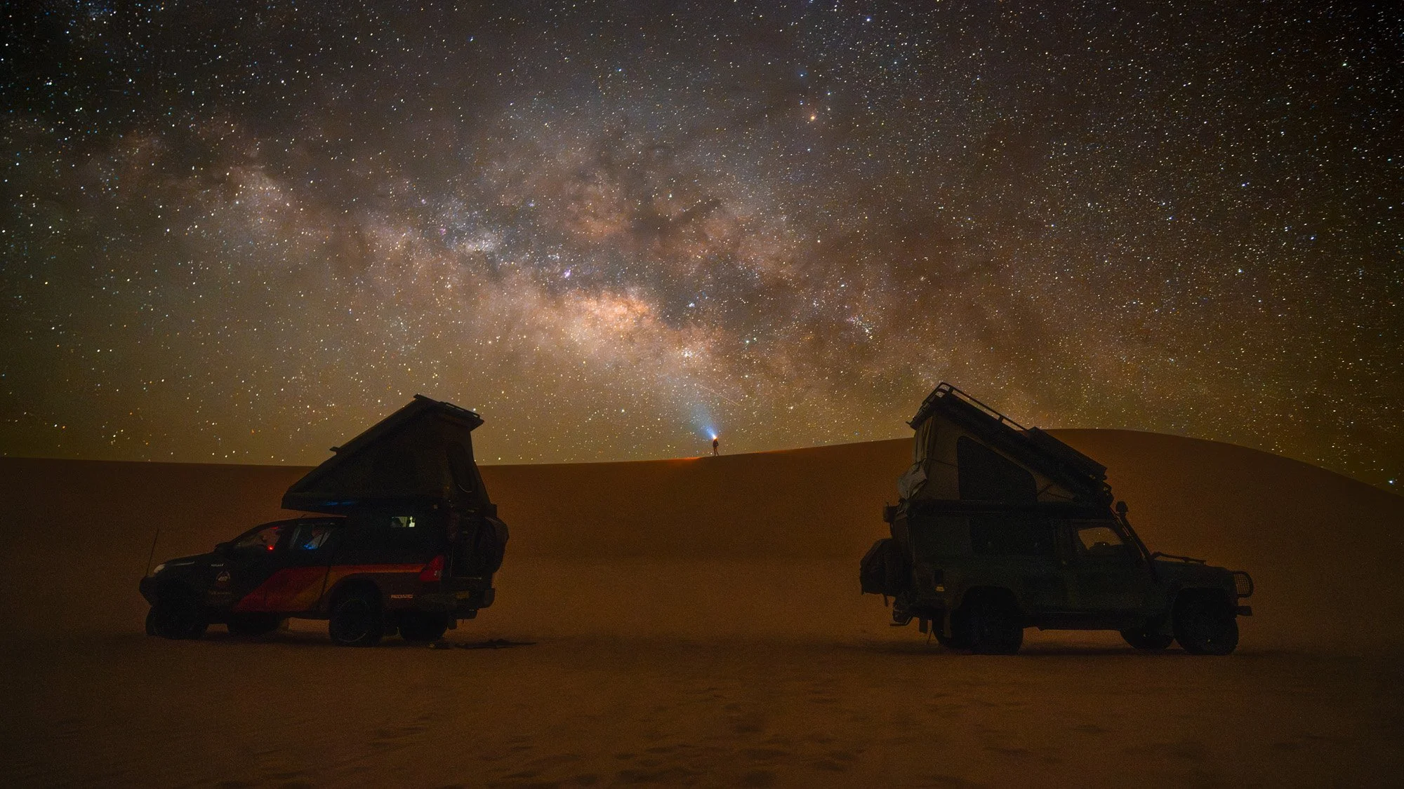 Two Land Rover Defenders parked under the brilliant core of the Milky Way galaxy in the Sahara Desert. The inclusion of the vehicles and a small light source provides a powerful sense of adventure and environmental scale.
