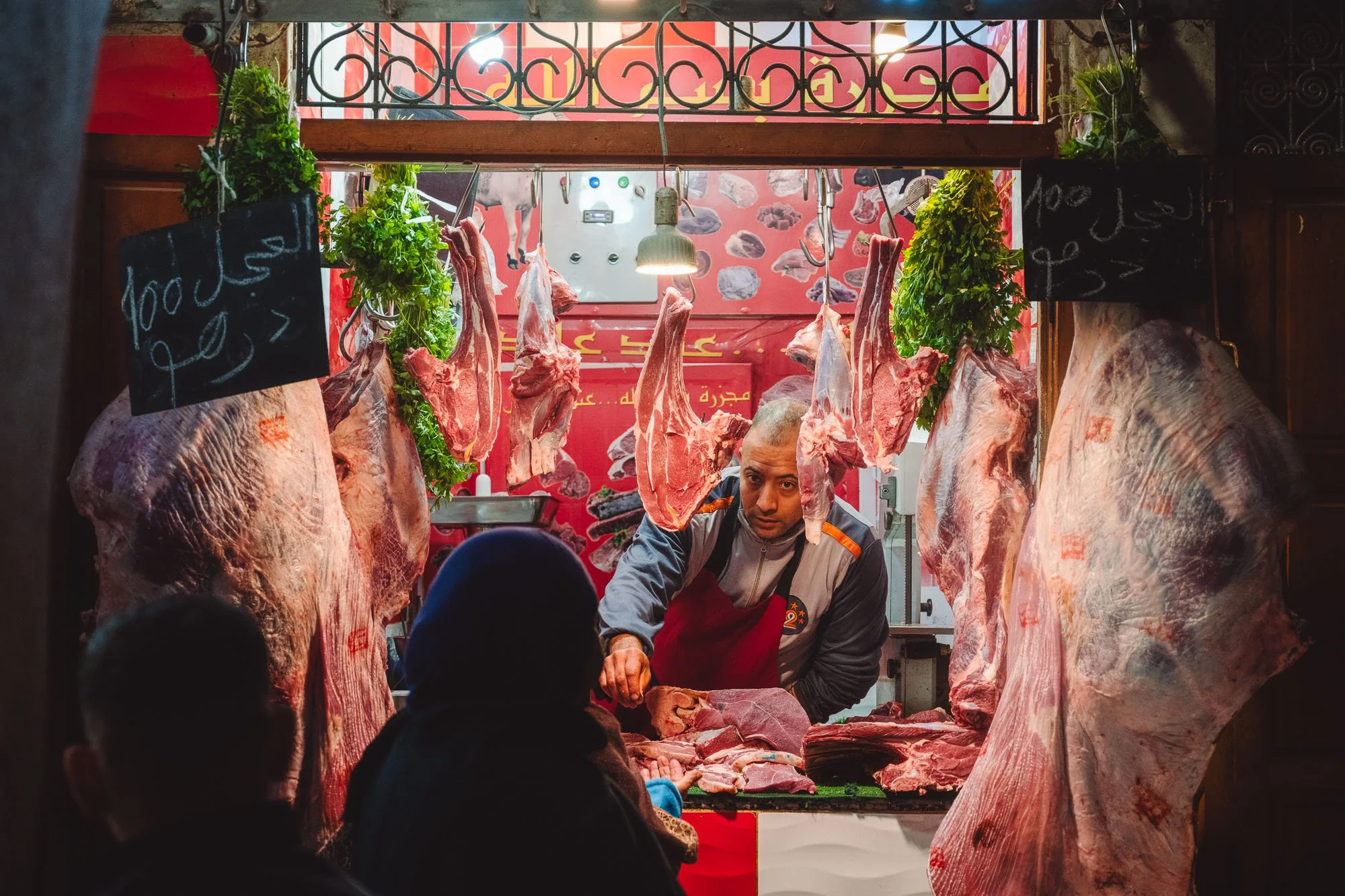 A butcher framed by hanging cuts of meat and chalkboards inside an atmospheric market stall in the Marrakech medina, Morocco