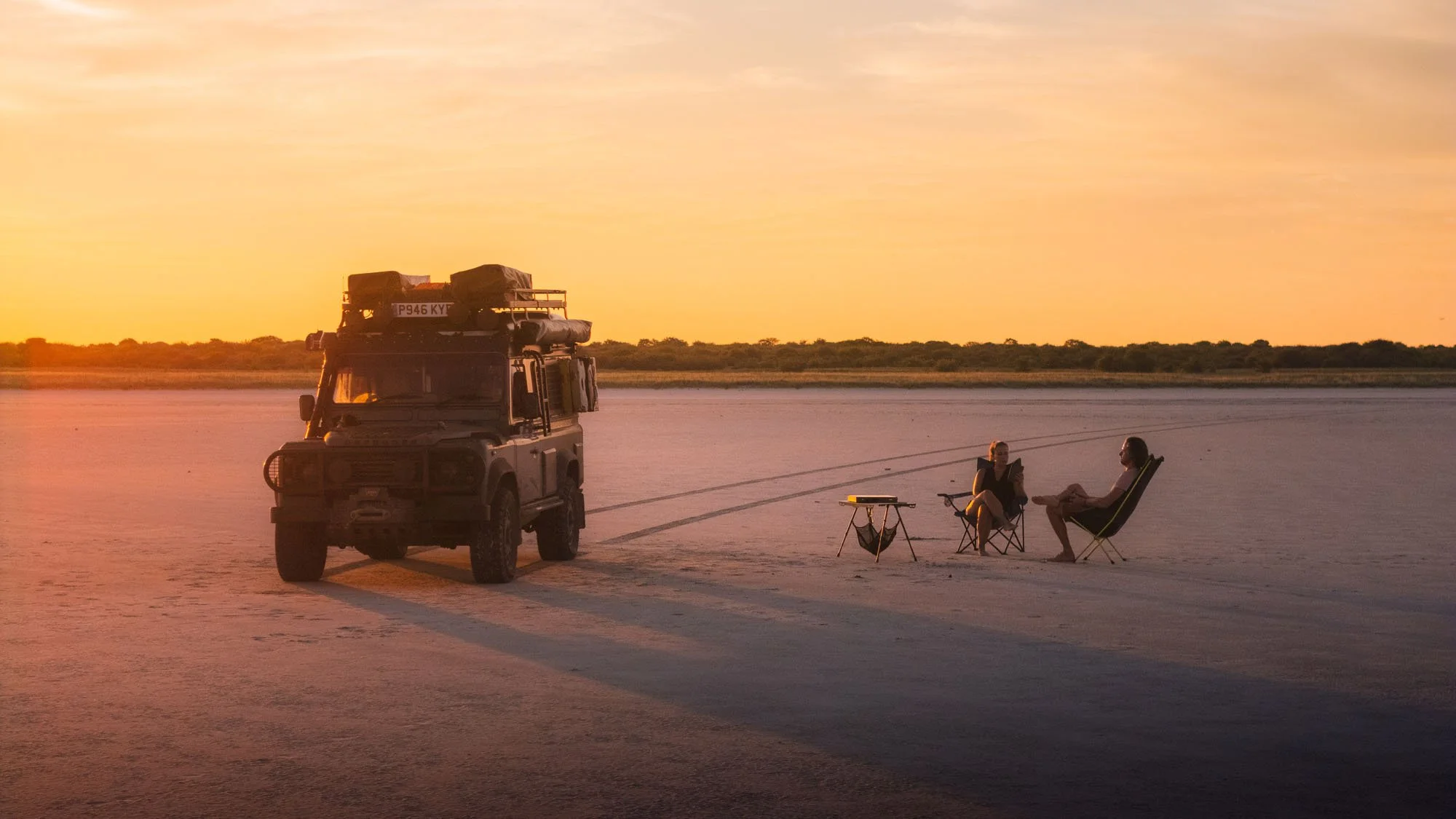 Two travelers sitting in folding camp chairs beside an overland Land Rover Defender on the vast, flat expanse of a Botswana salt pan at sunset.
