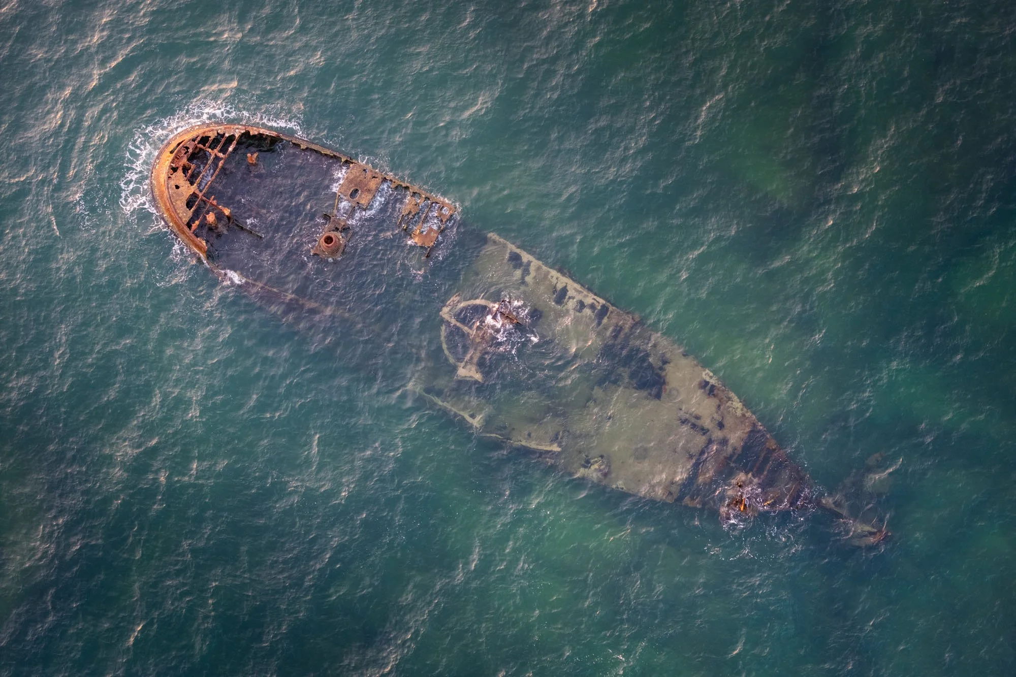 An aerial top-down view of a rusted, sunken shipwreck breaching the ocean surface. This image demonstrates Tip 5: 'Find the soul in the ugly,' using anomalous decay to trigger a dopamine spike.