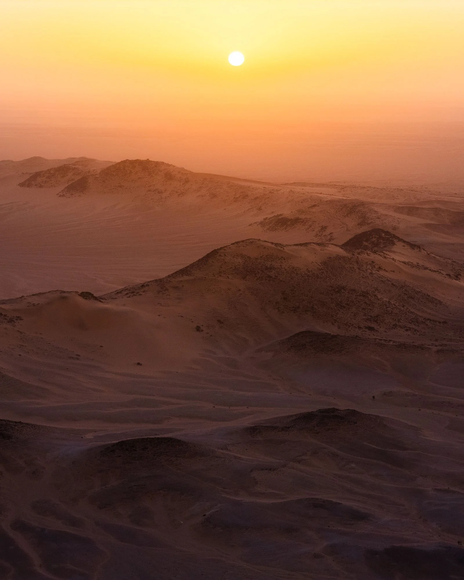 The sun setting through a thick haze over the vast, undulating desert dunes of the Tenoumer Crater in Mauritania, showing how visual ambiguity and lack of sharp horizons create perceptual friction