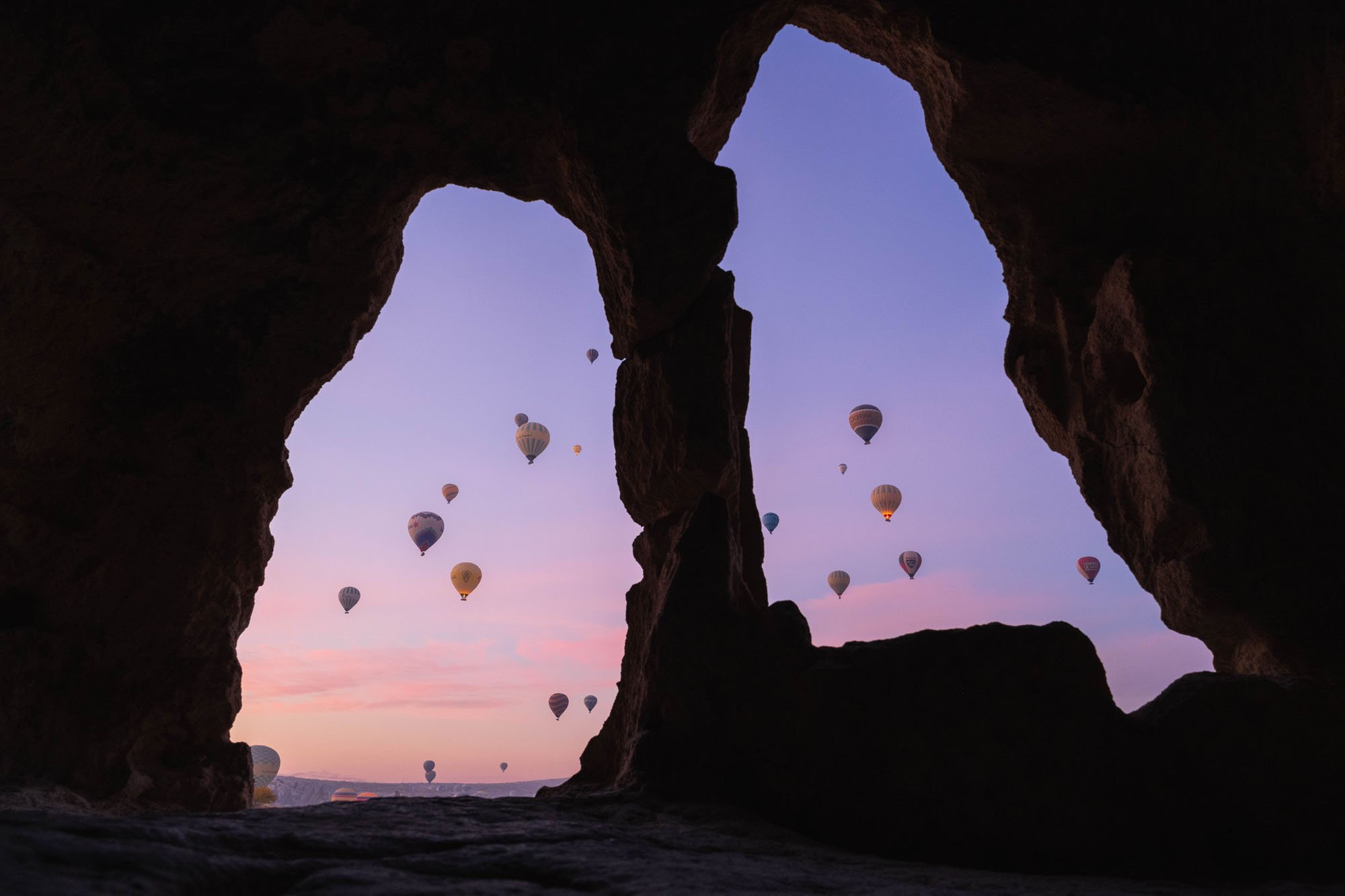 Hot air balloons drifting across a violet dawn sky as seen through the dark mouth of an ancient cave in Cappadocia, demonstrating the visual relationship between interior and exterior spaces