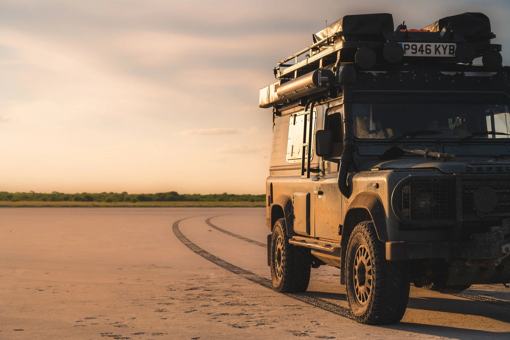 An expedition-equipped Land Rover Defender 110 parked on the vast, flat expanse of the Makgadikgadi Salt Pan in Botswana at sunset, with curving tyre tracks in the foreground