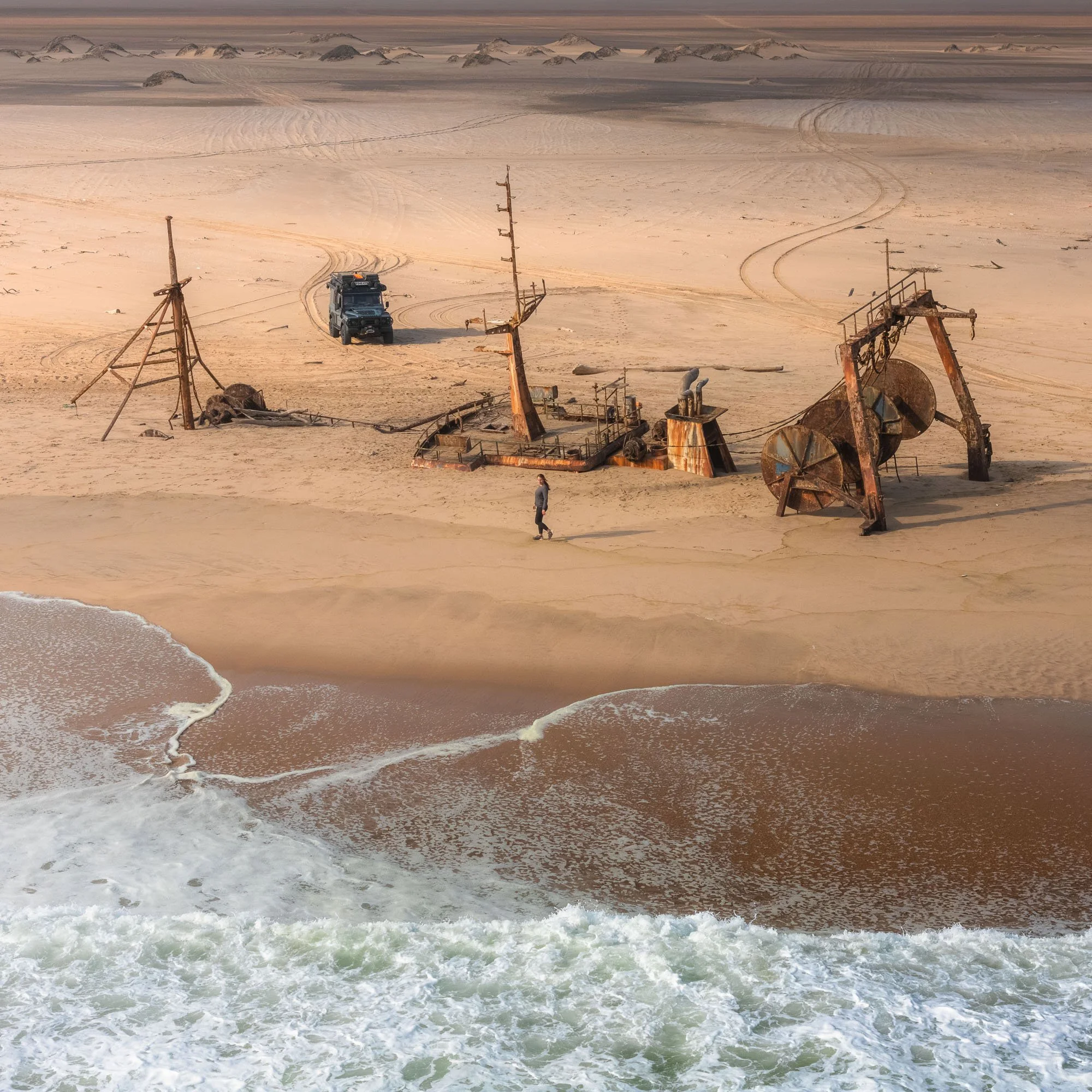 High-angle view of a Land Rover Defender and a lone traveler standing beside a massive rusted coastal shipwreck on a sandy beach in Angola.