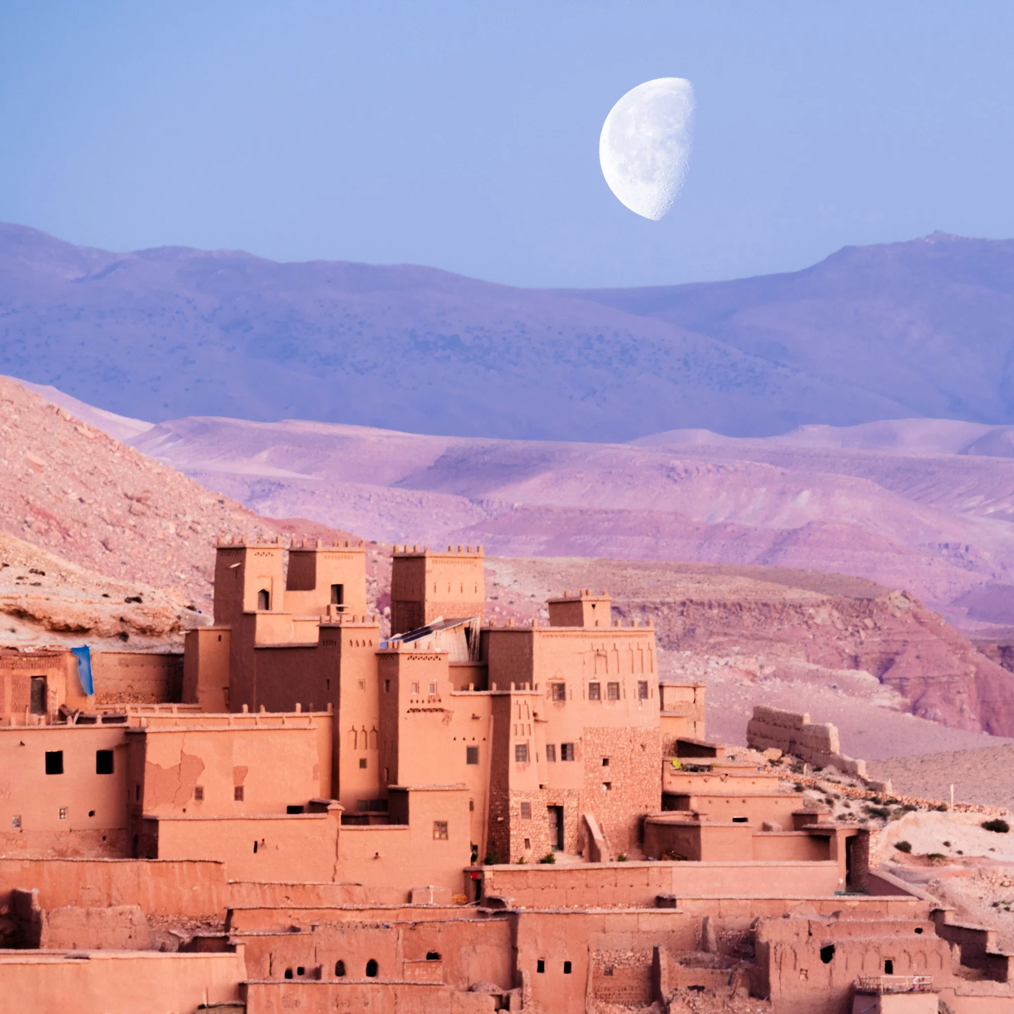 The ancient clay city of Ait Benhaddou, Morocco, set against purple mountains beneath a massive moon. The layering of the mountains utilizes atmospheric perspective to create a deep psychological sense of place.