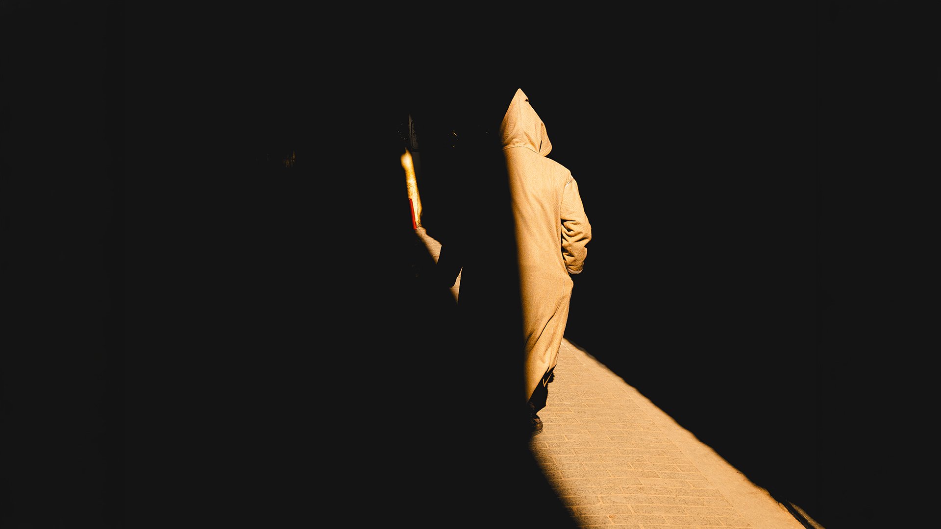 A hooded figure in a traditional djellaba walking down a narrow diagonal shaft of sunlight cutting through deep black shadows in a Moroccan alleyway.