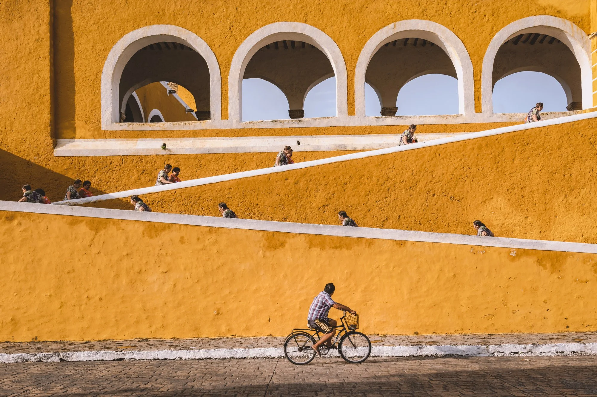 A man riding a bicycle in front of a vibrant yellow building with arched windows. Several people are seated on the building's sloped white ledges, enjoying the sunny day.