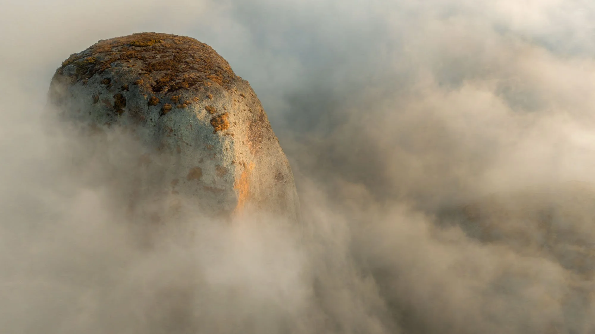 A massive, isolated rock formation jutting out of thick, swirling fog at Pedras Negras de Pungo Andongo in Angola, leaving an unresolved visual puzzle that commands human attention