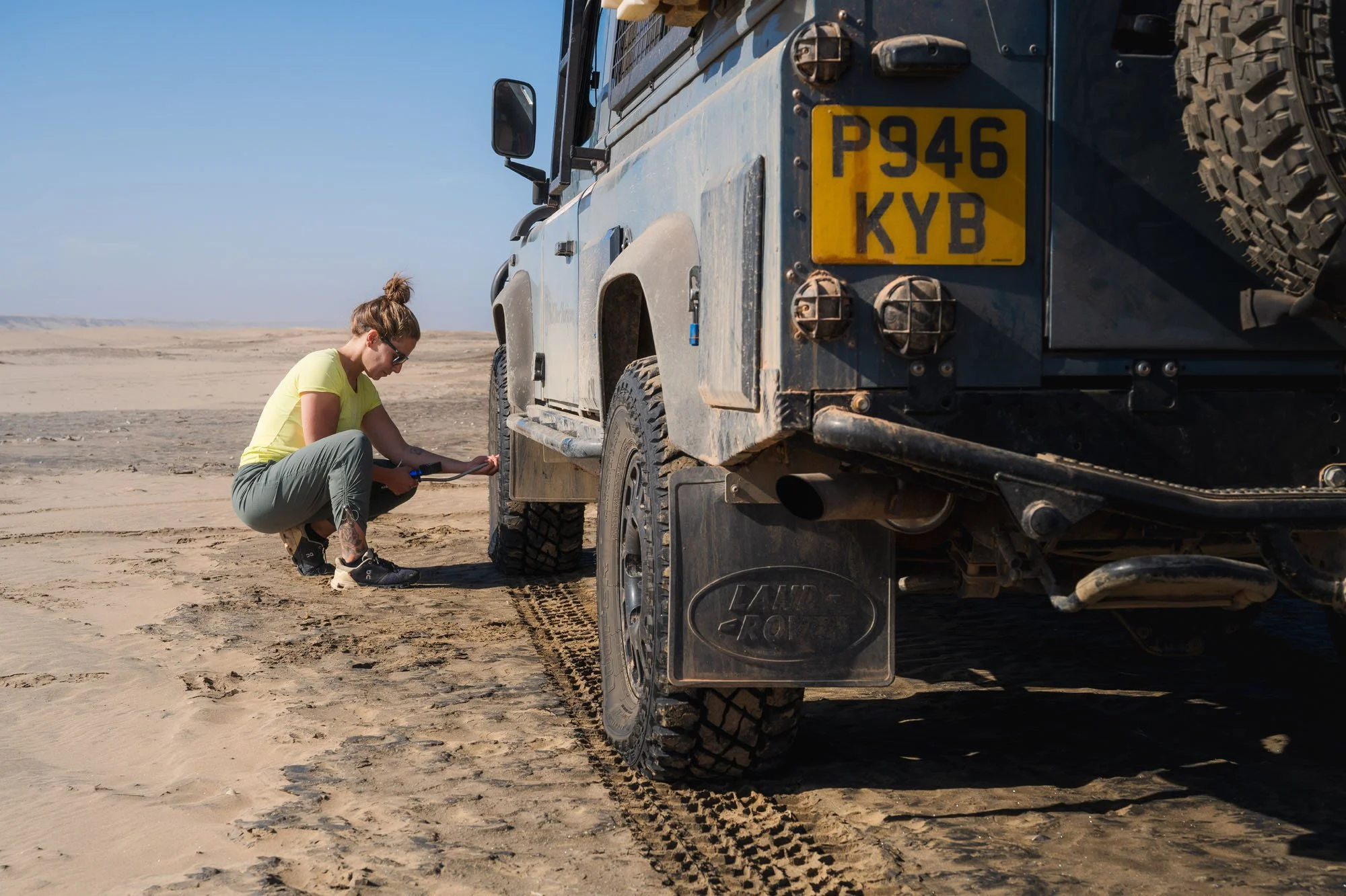 A traveler kneeling to air down the tyres on the rear of an overland Land Rover Defender 110 on a sandy track in Africa