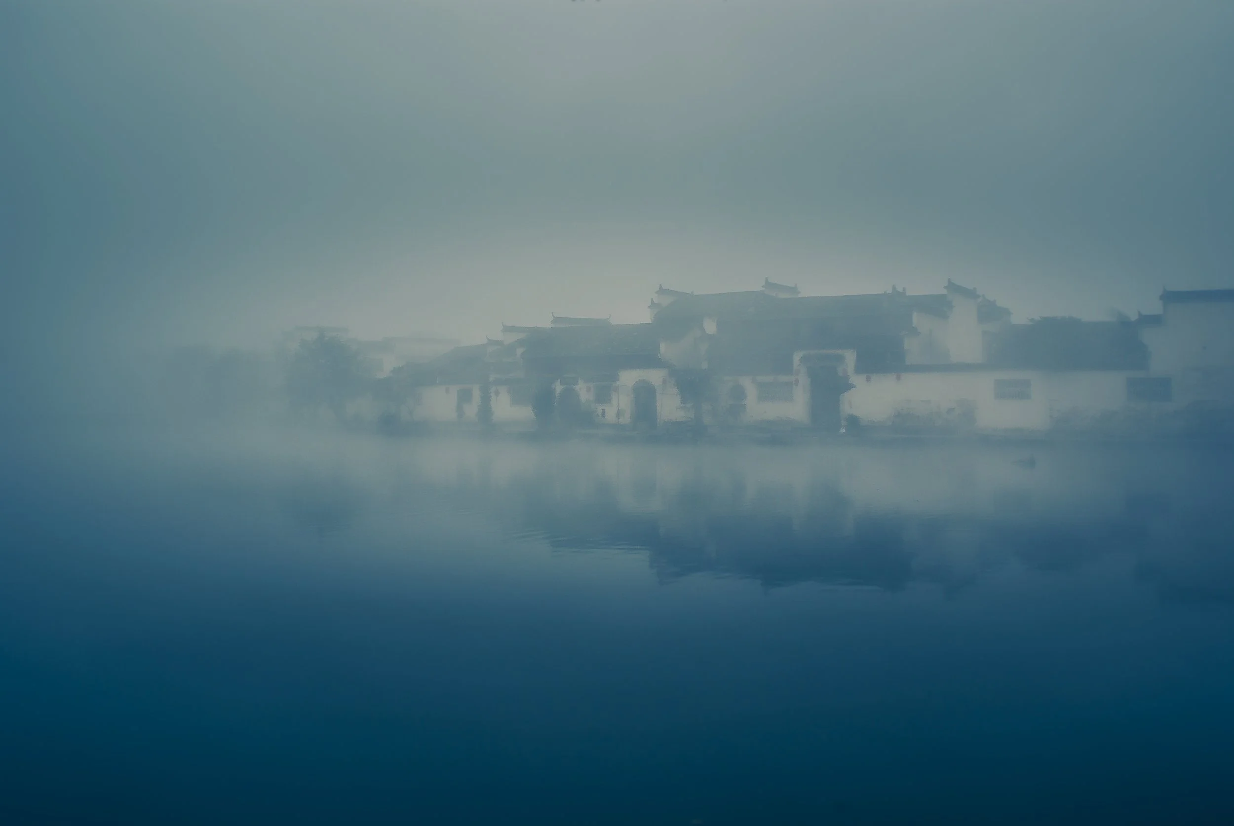 A foggy scene of traditional Asian buildings reflected on a body of water.
