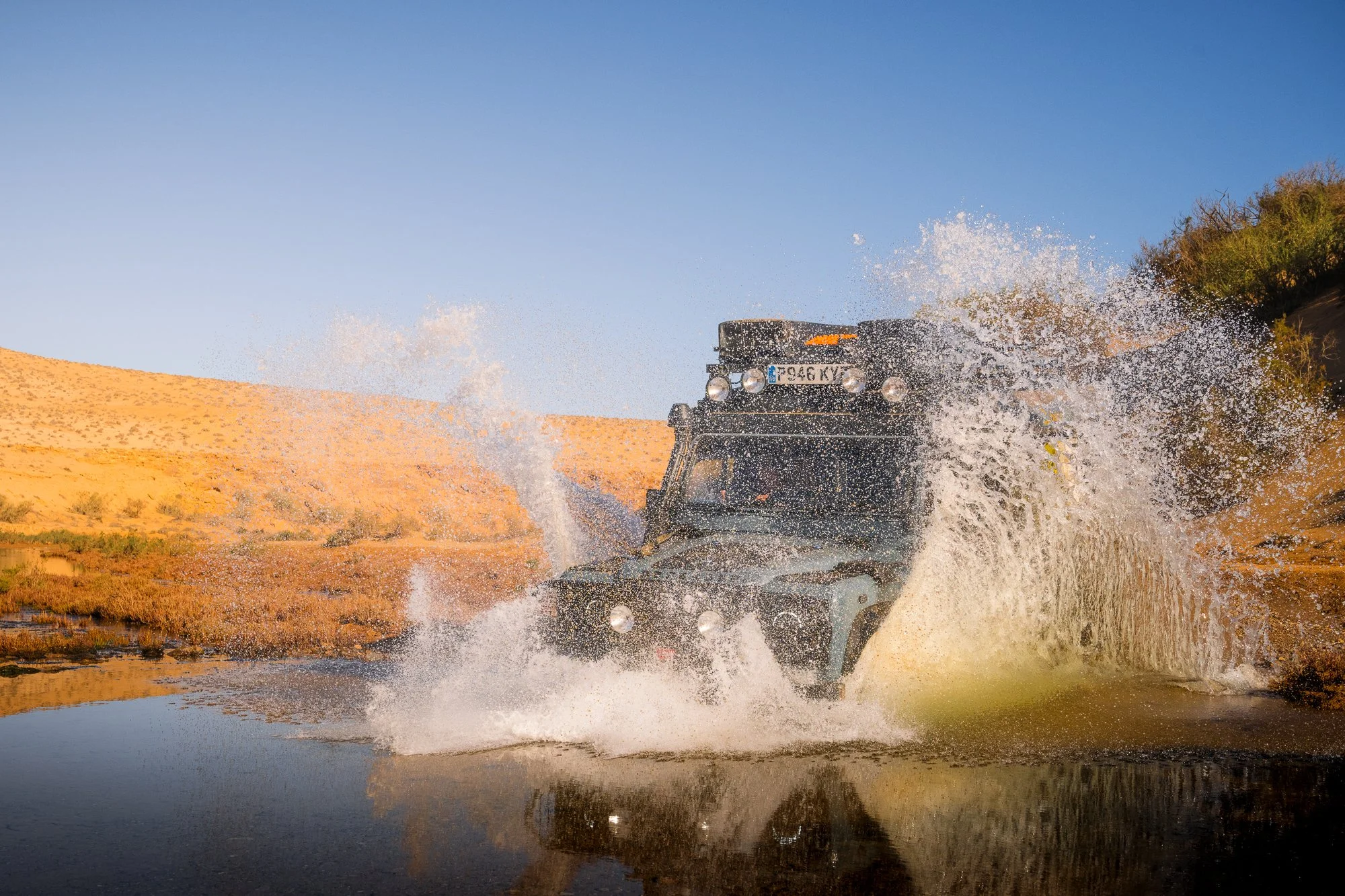 A black off-road vehicle driving through water, creating a splash in a desert landscape with hills, clear sky, and some vegetation.