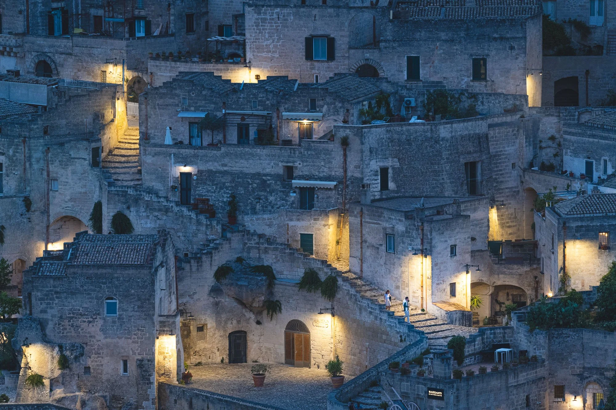 A panoramic twilight view of the historic stone buildings and staircases of Matera, Italy, illuminated by warm glowing streetlamps