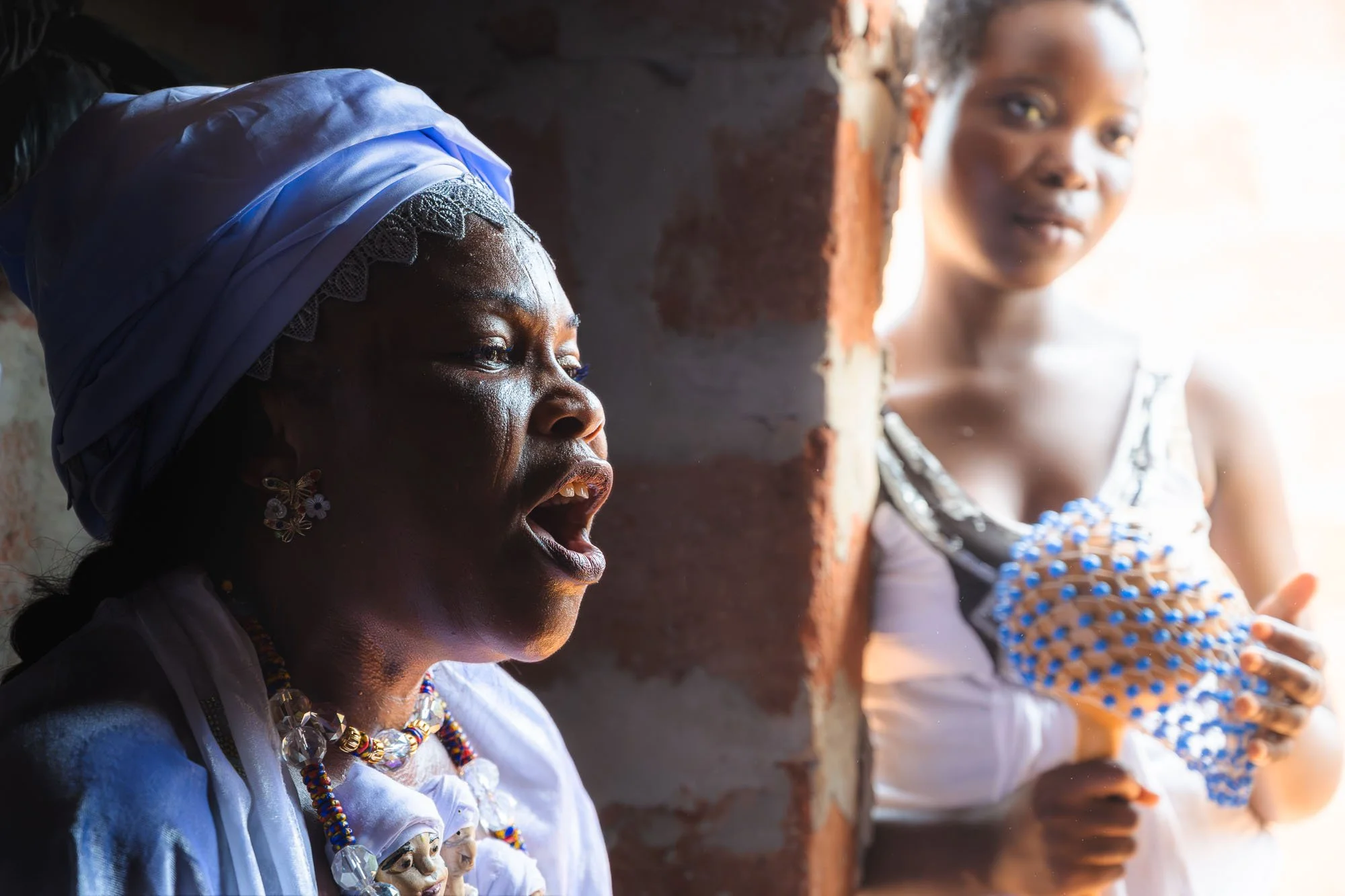 A Mami Wata initiate chanting passionately with eyes closed while holding beaded gourd rattles inside an intimate shrine in Benin