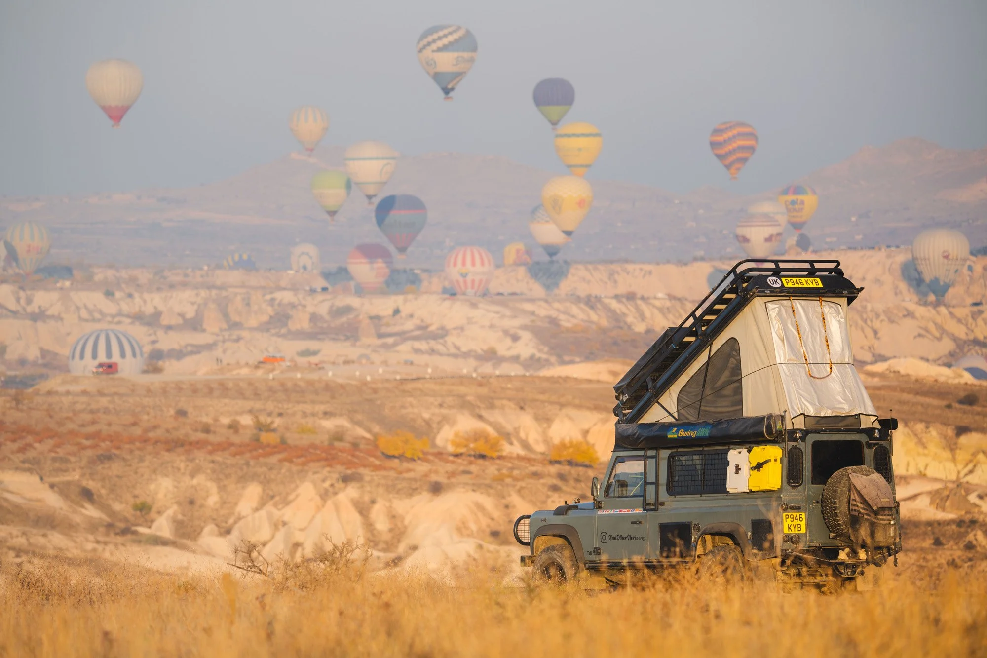 A Land Rover Defender parked in a golden grassy field with dozens of hot air balloons filling the background in Cappadocia. The layering of subjects from foreground to background creates a deep sense of narrative context.