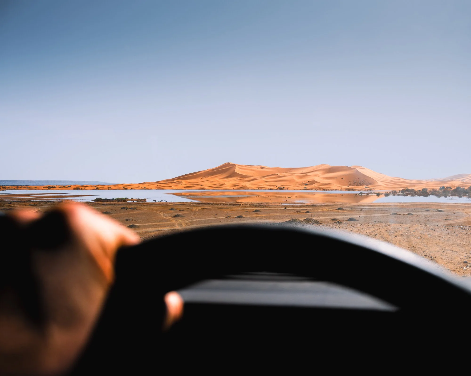 A POV shot from the driver's seat of a Land Rover Defender looking toward a distant sand dune. This is a practical example of amodal completion, where showing only the hand and wheel prompts the brain to fill in the rest of the expedition's scale.
