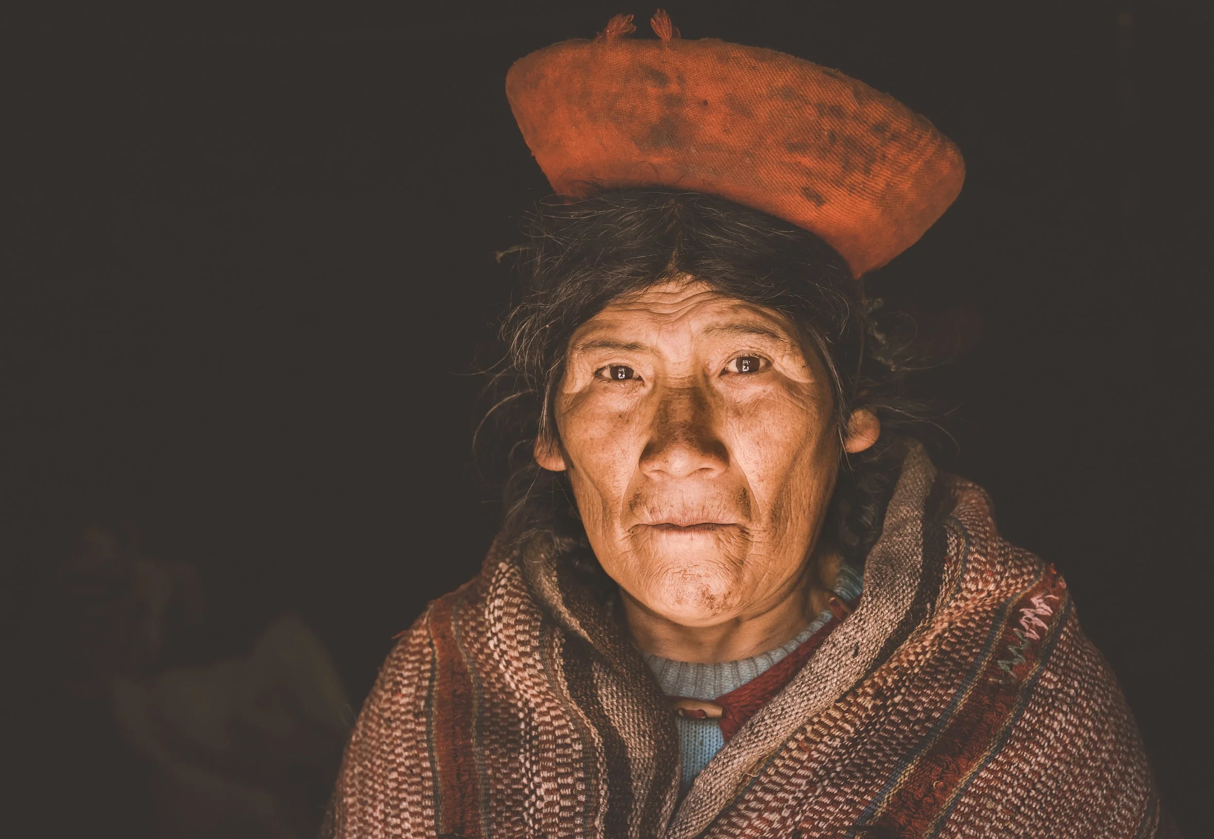 Portrait of an elderly woman with deeply lined face, wearing a traditional woven shawl and a hat resembling a large, round, orange fruit, against a dark background.