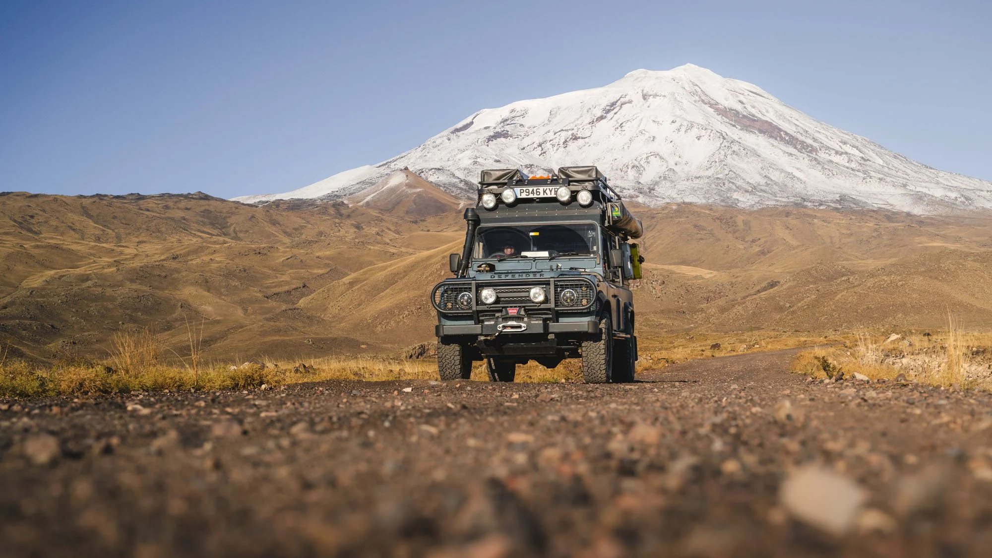 A 1997 Land Rover Defender parked on a dusty track with the snow-capped peak of Mount Ararat in the background, showing a classic photography adventure schema.