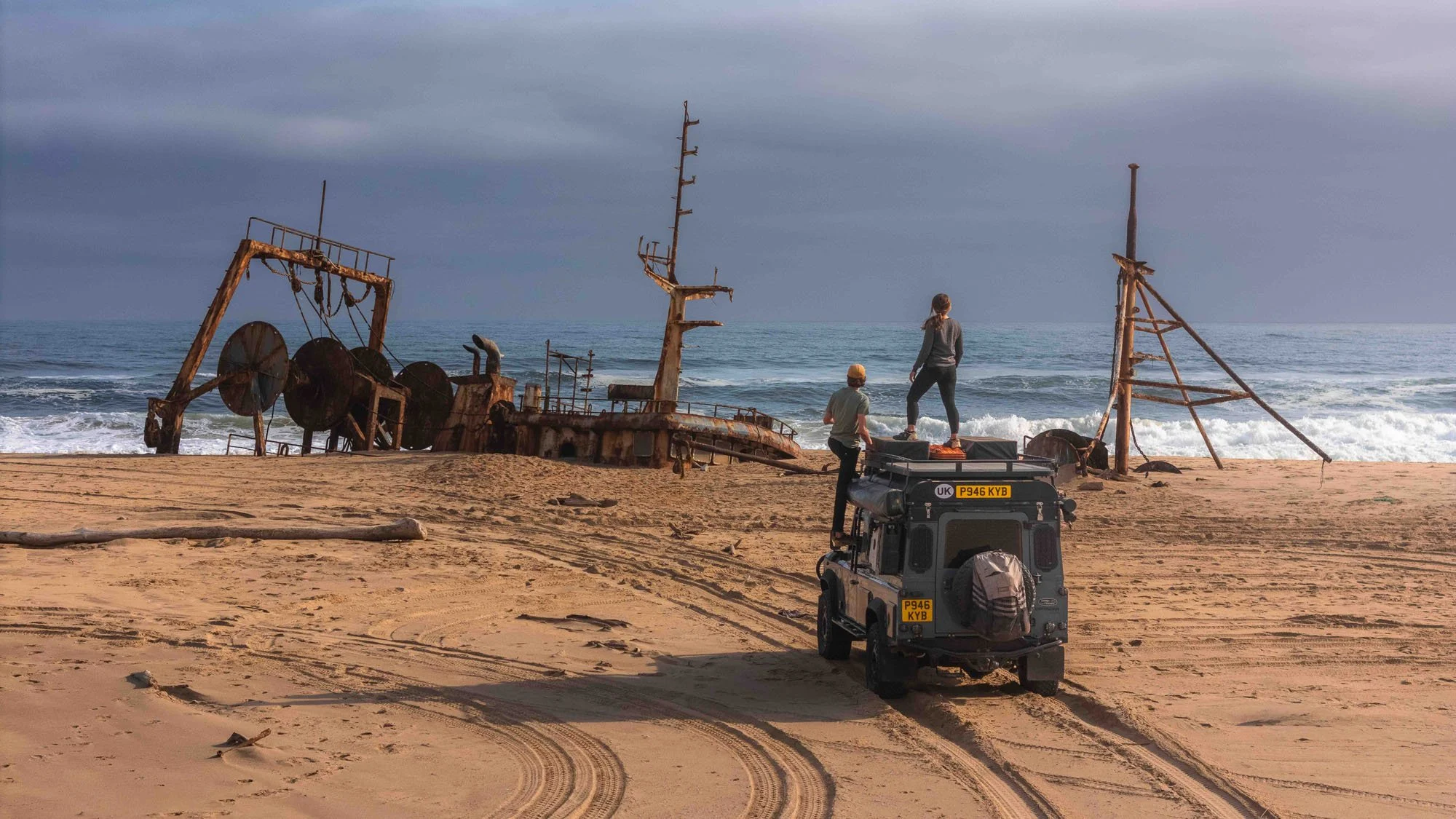 Two people stand atop a Land Rover Defender on the desert shores of Angola, looking at the rusted Vanessa shipwreck. The photograph is a record of an encounter, capturing the raw reality of the world over technical perfection.
