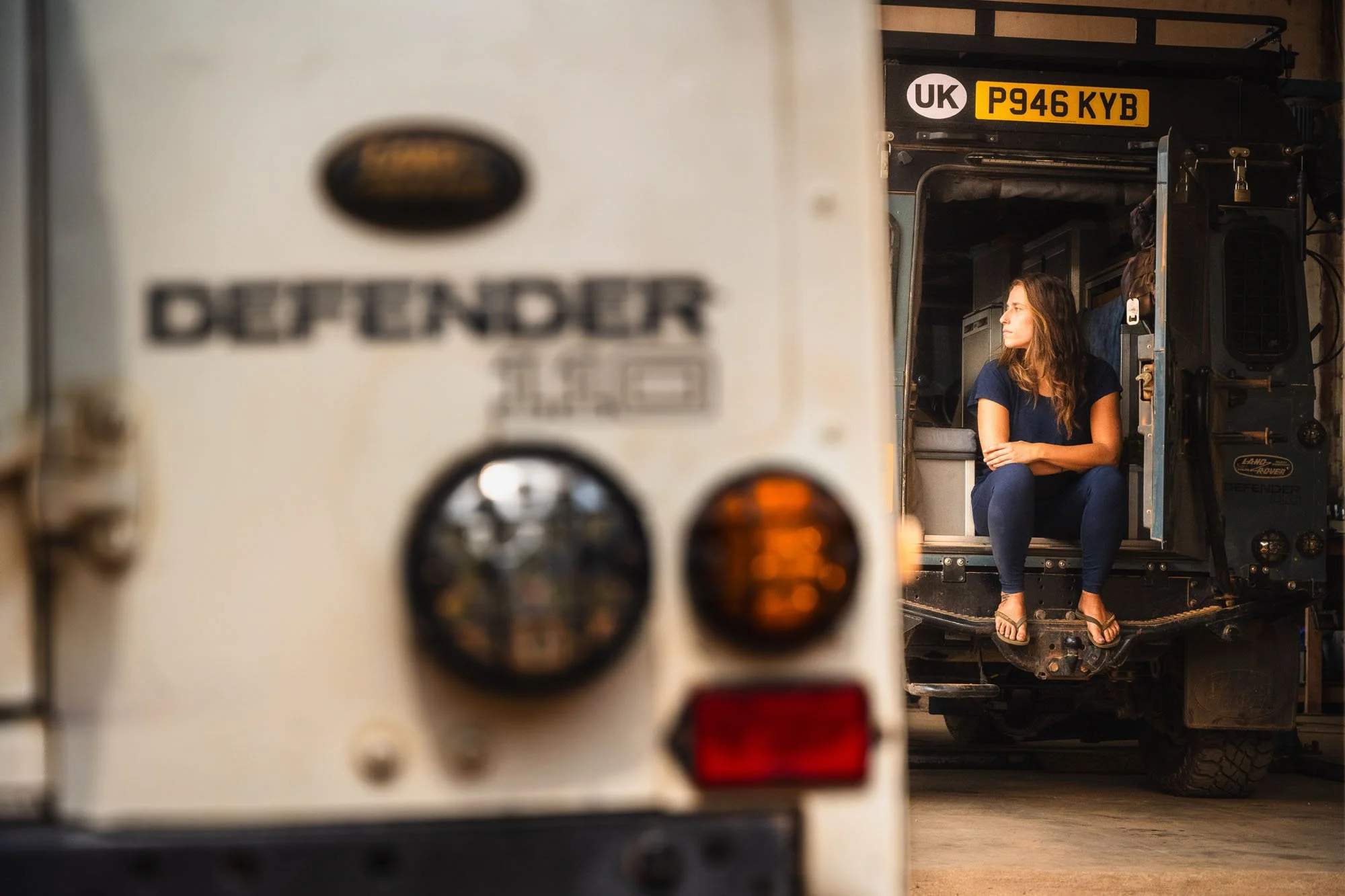 A woman sitting thoughtfully in the open rear door of an expedition-equipped Land Rover Defender during an overland trip in Angola.