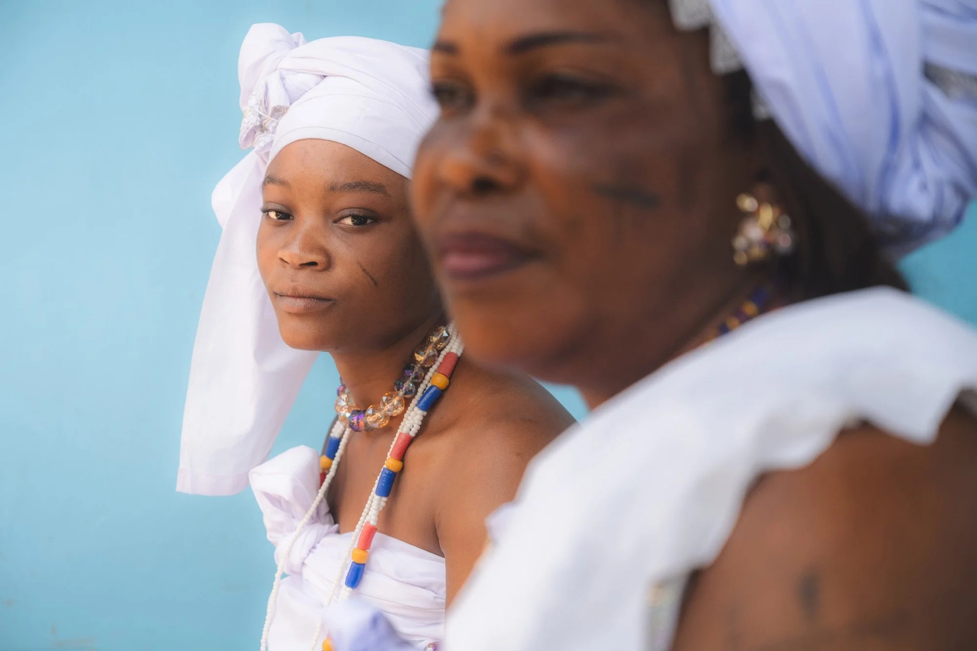 A young woman wearing traditional white clothing and a head wrap, with jewelry, looking at an older woman dressed similarly with head wrap, earrings, and jewelry, both against a blue background.