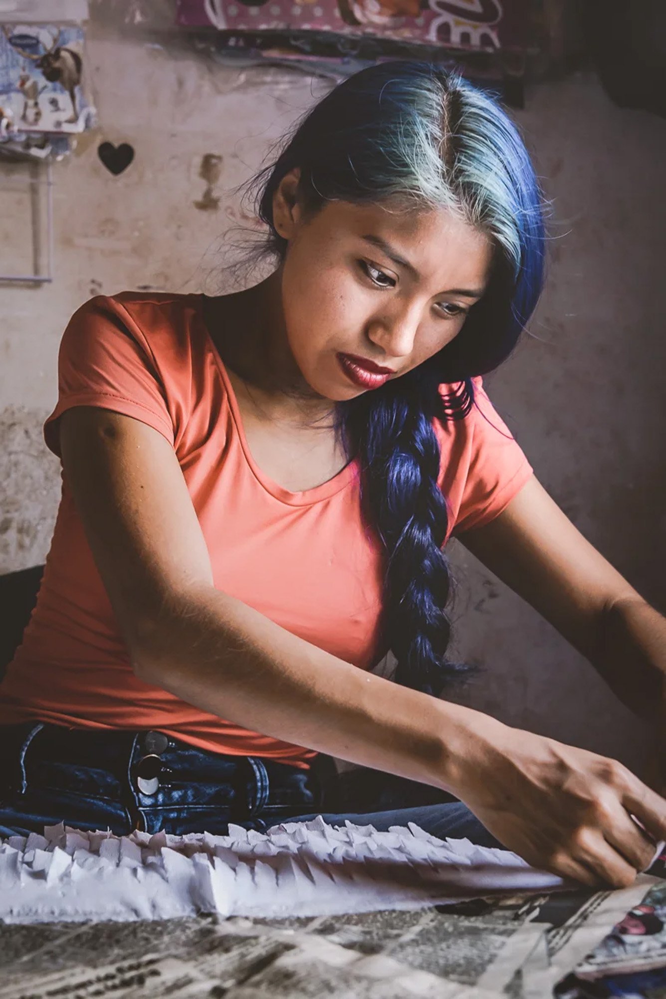 A soft portrait of a young woman with blue hair working in Mexico. The gentle use of chiaroscuro lighting and her downward gaze utilize 'the art of withholding' to create a sense of intimacy and quiet focus.