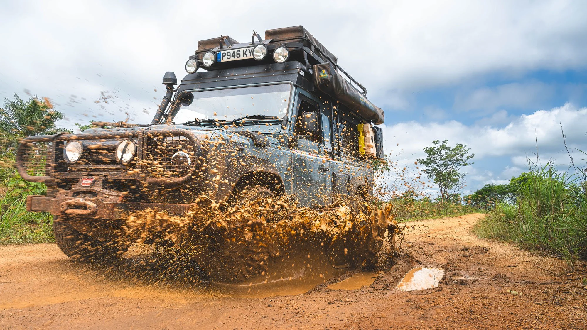 An expedition-equipped Land Rover Defender 110 ploughing through deep red mud and water on a remote road during the wet season in Benin.