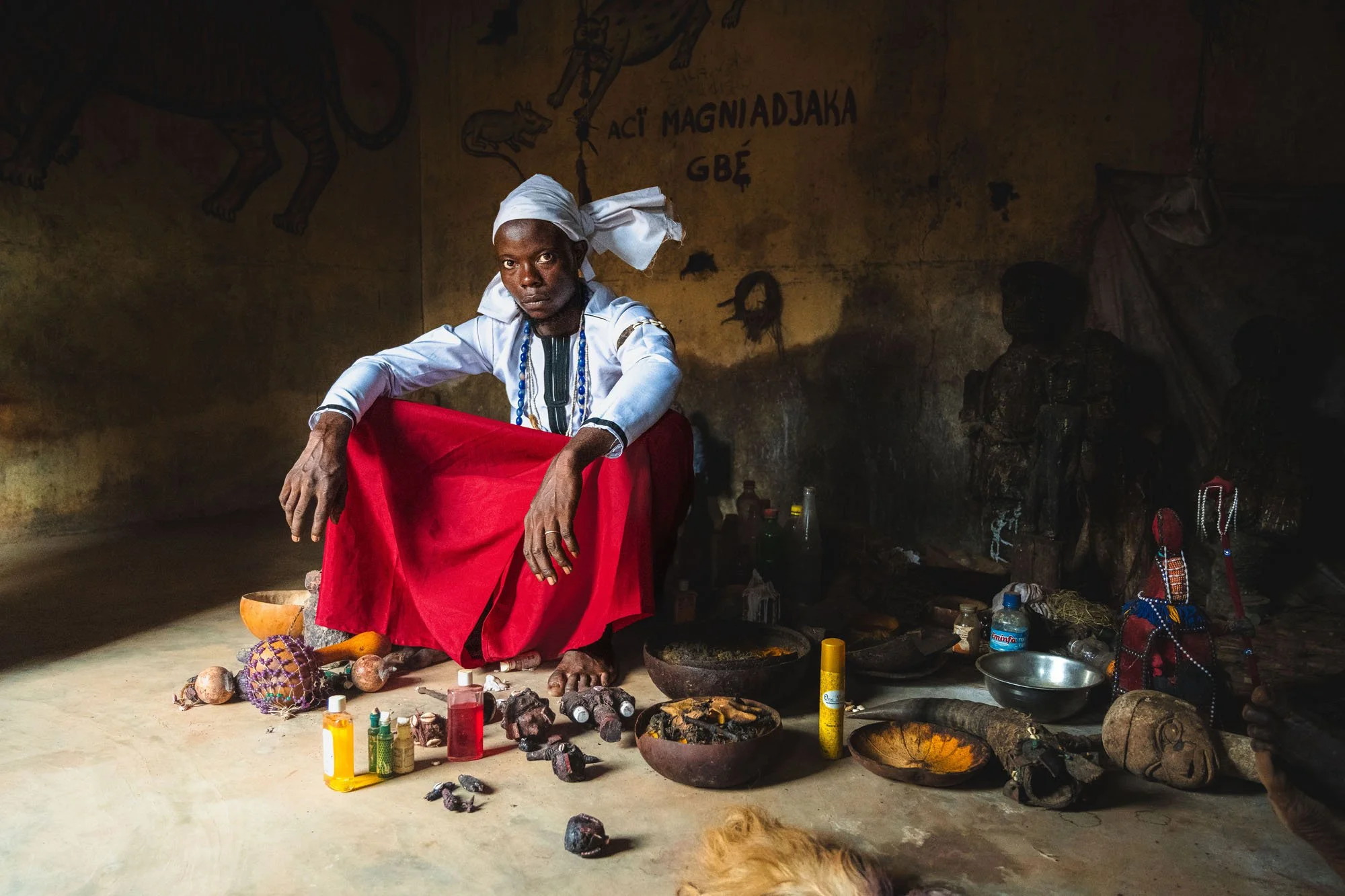 A Voodoo priest in Benin sits cross-legged in a shadow-heavy shrine, surrounded by ritual objects. His direct stare forces the viewer to engage with the authentic weight of the scene.