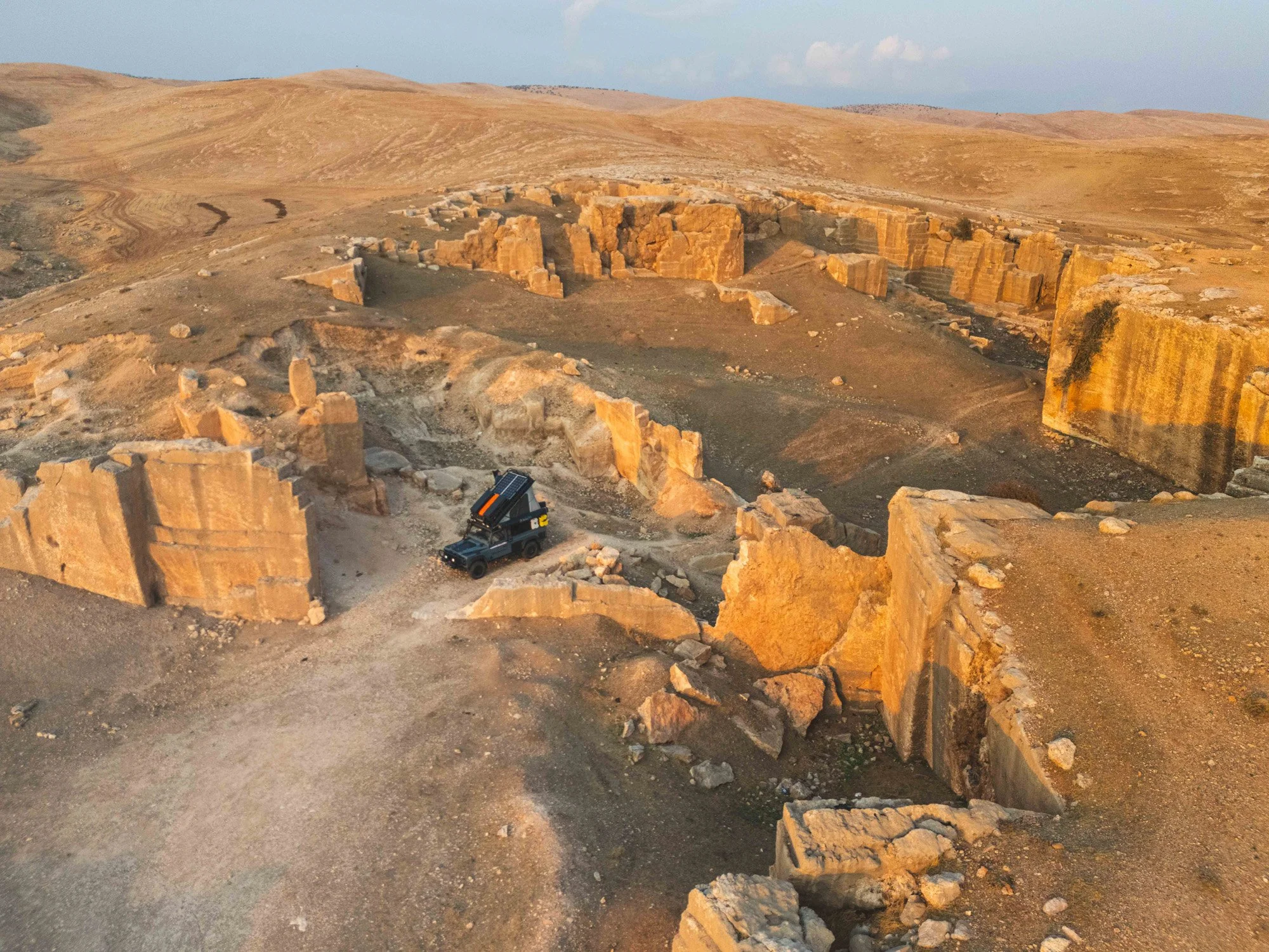 A Land Rover Defender parked amidst the sprawling Roman-era rock-cut quarry of Dara, Mesopotamia. The juxtaposition of the modern expedition vehicle and the ancient ruins creates a powerful visual anomaly.