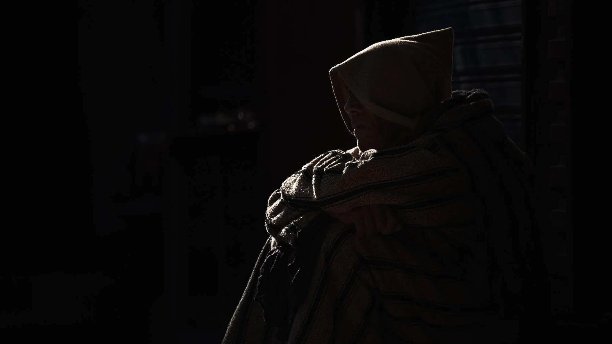 A dramatic, low-key portrait of a cowled man in a Moroccan doorway swallowed by deep shadows, using the psychology of amodal completion to hide identity and engage the viewer