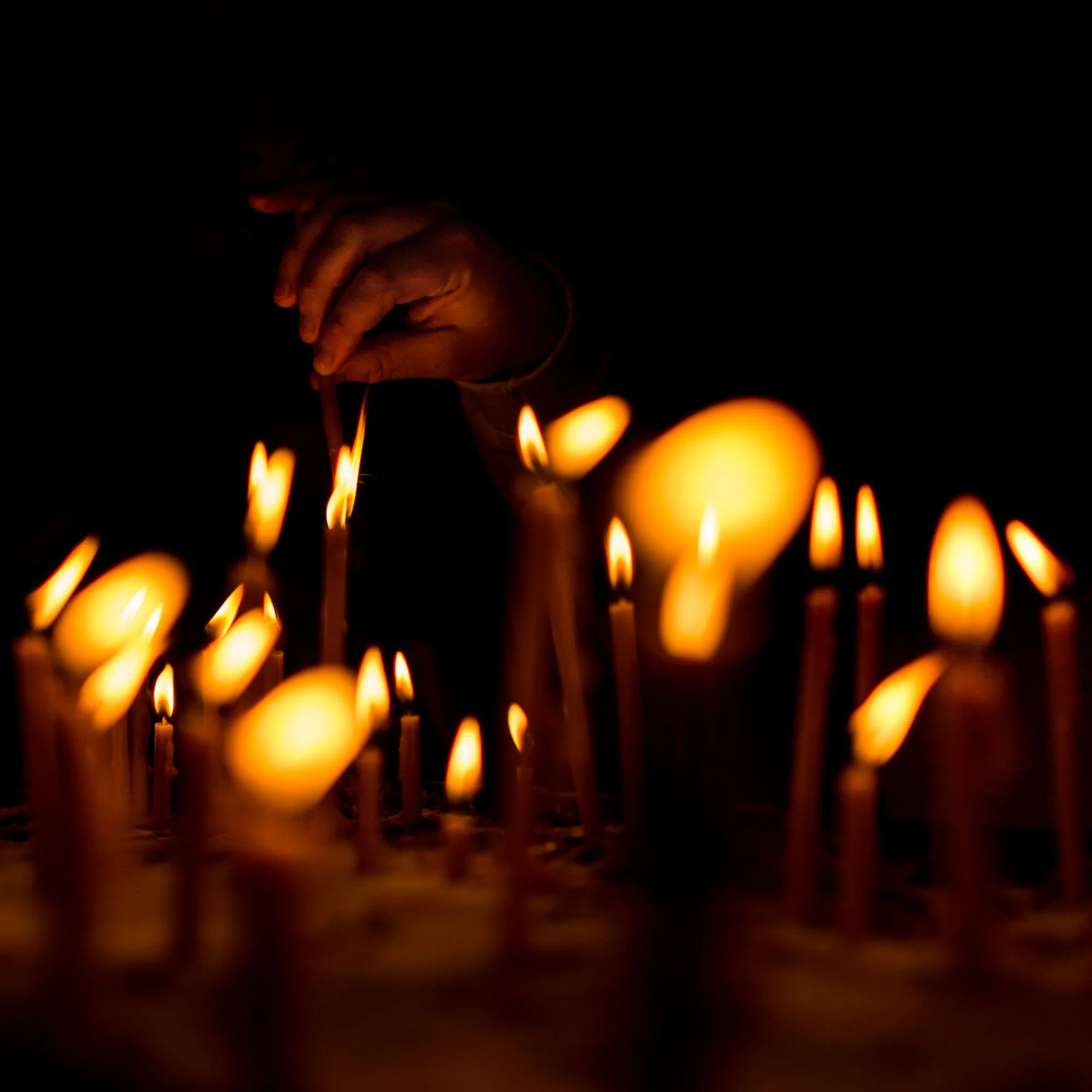 A close-up of a hand lighting thin candles in an atmospheric room with glowing golden flames cutting through the deep black background in Vienna.