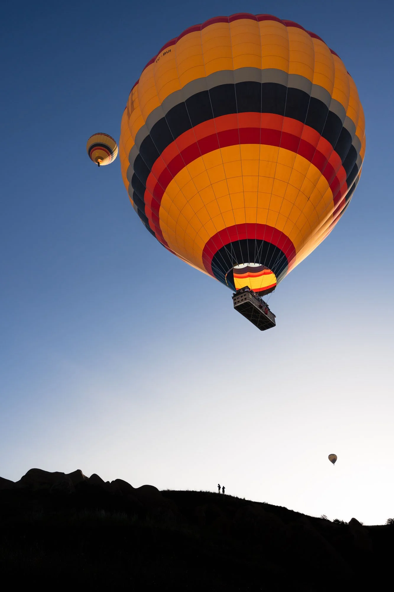 A colossal yellow and orange hot air balloon hovering directly over a black silhouette ridge with two tiny human figures in Cappadocia, Türkiye.