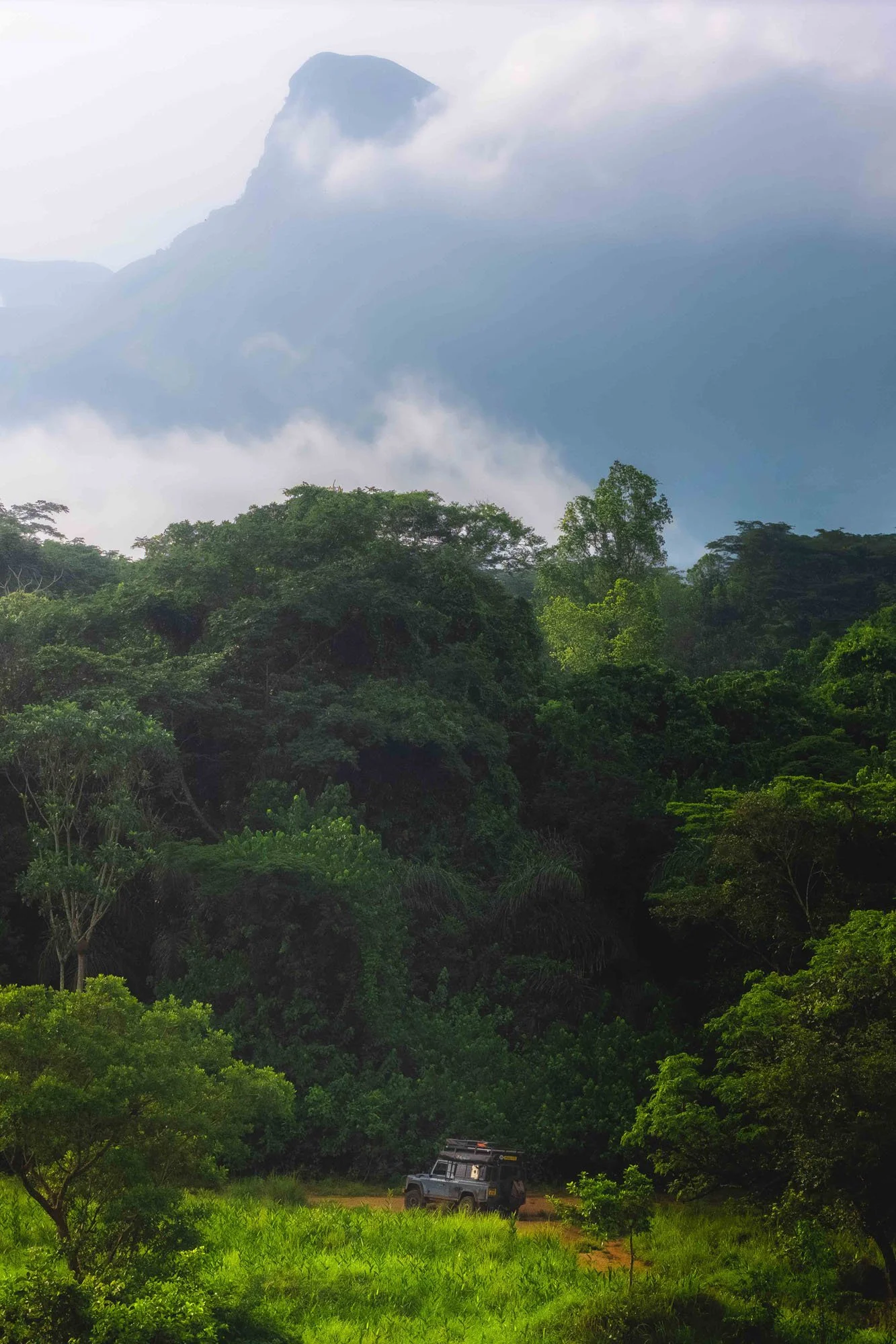 A tiny Land Rover Defender drives through a massive, lush jungle valley in the Guinea Highlands under heavy mist. The extreme scale highlights human vulnerability and the importance of remaining present in the sensory richness of the moment.