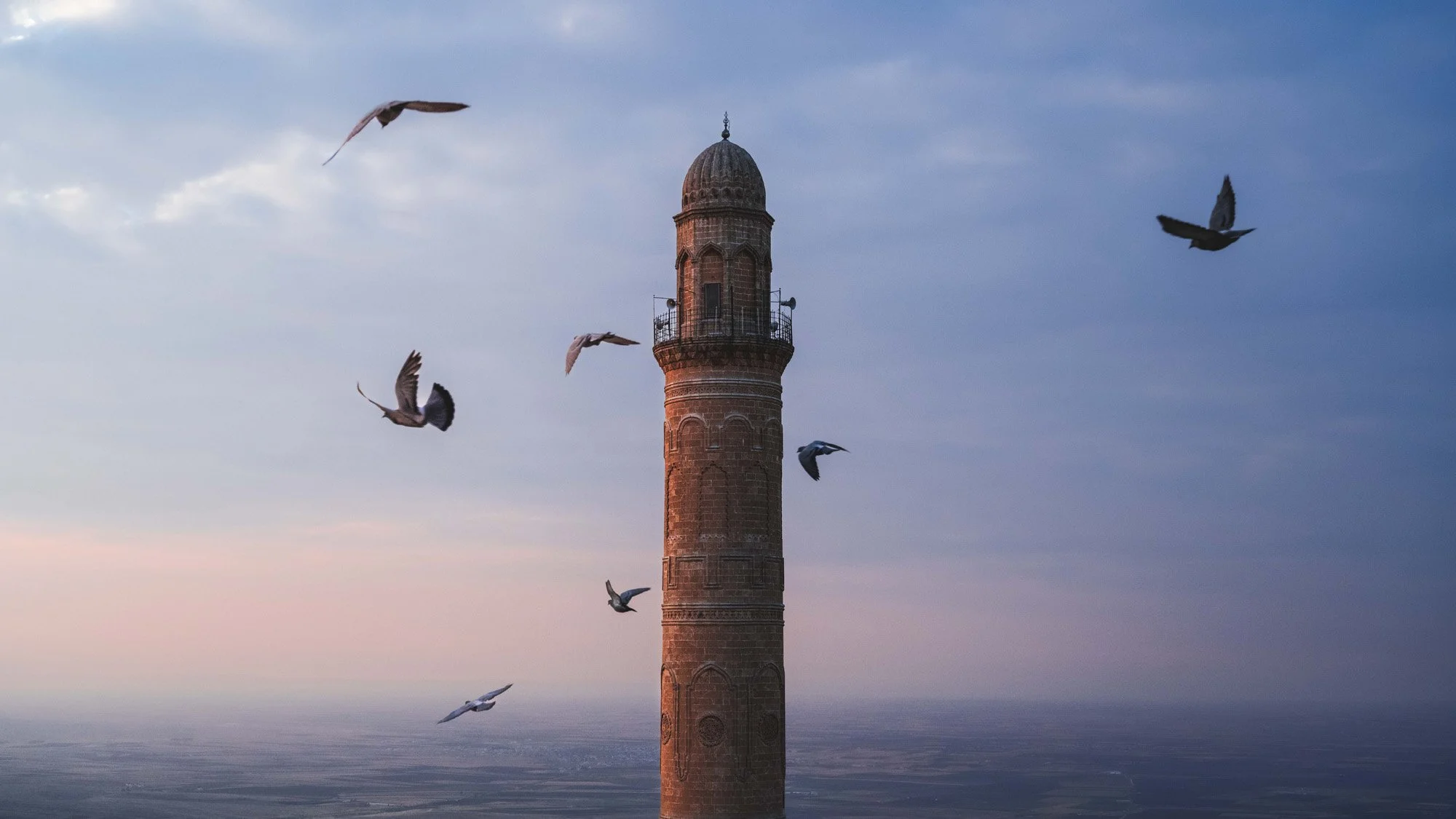 An ancient stone minaret in Mardin, Türkiye, surrounded by a dramatic flock of pigeons in mid-flight against a dusky evening sky.