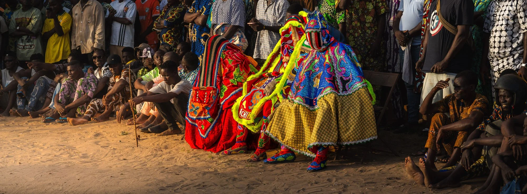 Three ornate Egungun figures seated quietly alongside village elders in the shade during a traditional ceremony in Ouidah, Benin