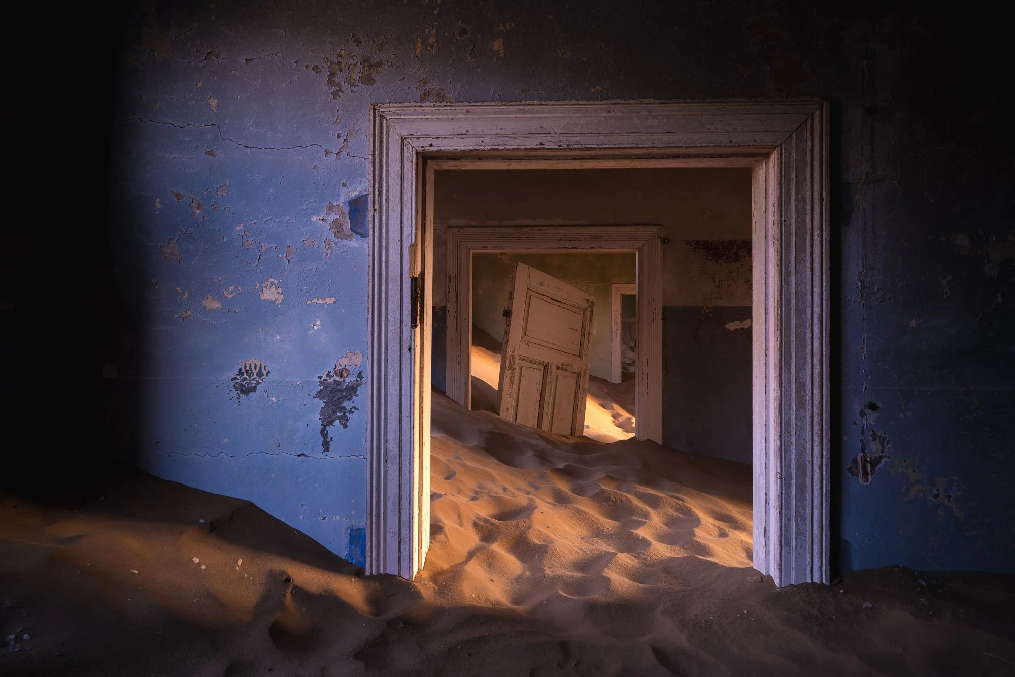 Desert sand dunes spilling through an open doorway into an abandoned, decaying blue bedroom in the ghost town of Kolmanskop, Namibia, lit by soft morning sunlight