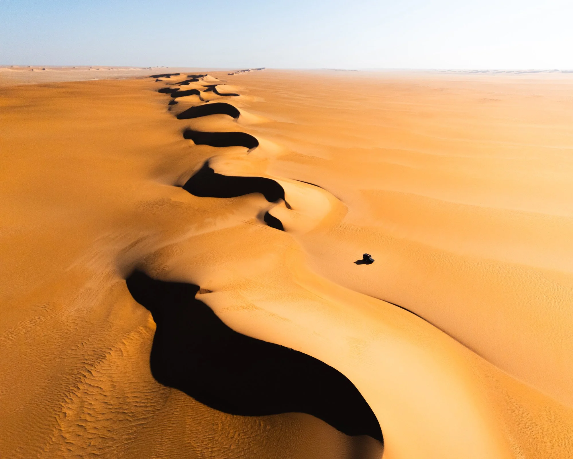 A vast desert with golden sand dunes and deep black shadows cast by the dunes, extending into the horizon under a clear sky.